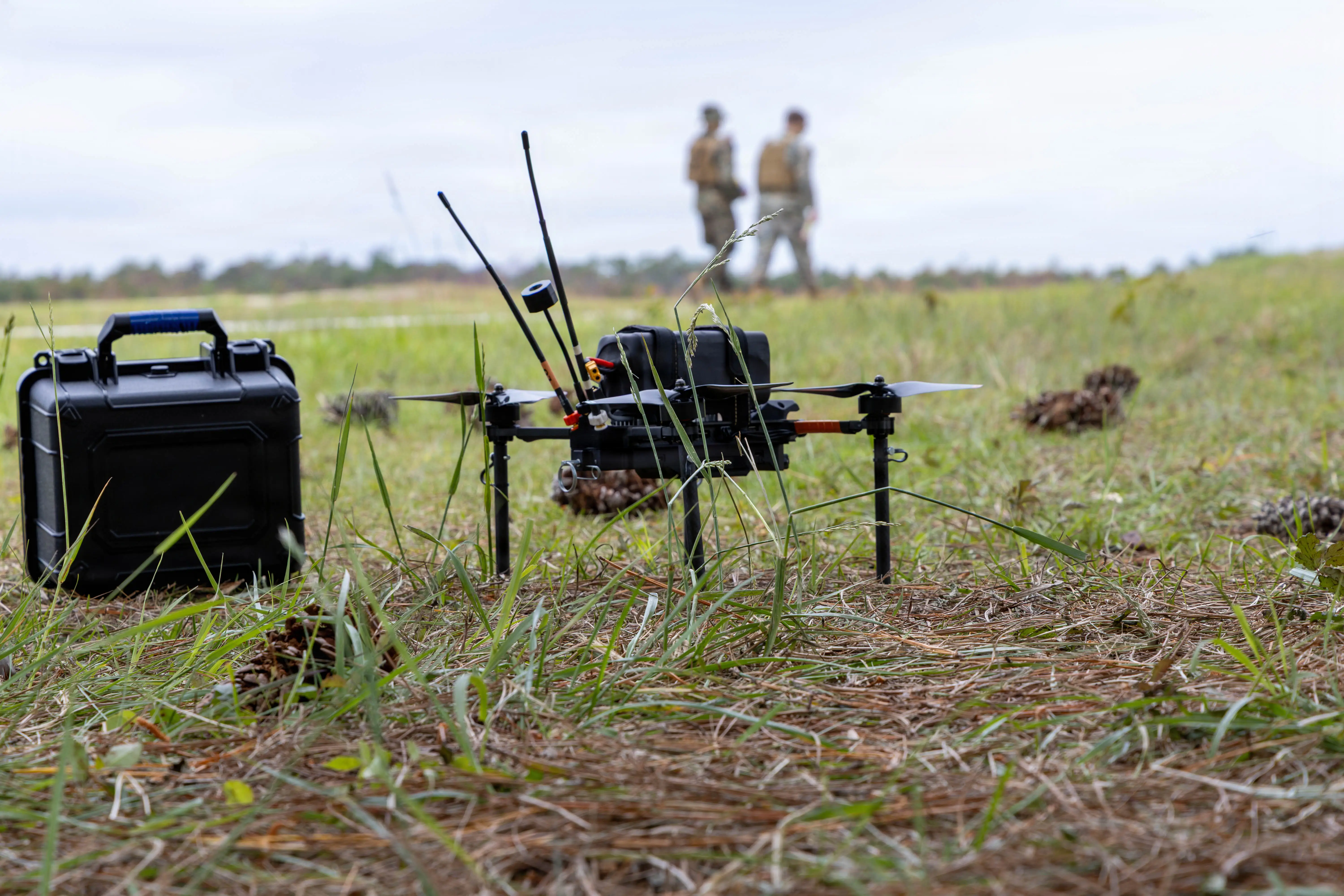 A small drone sits on the grass with two Marines walking in the background.