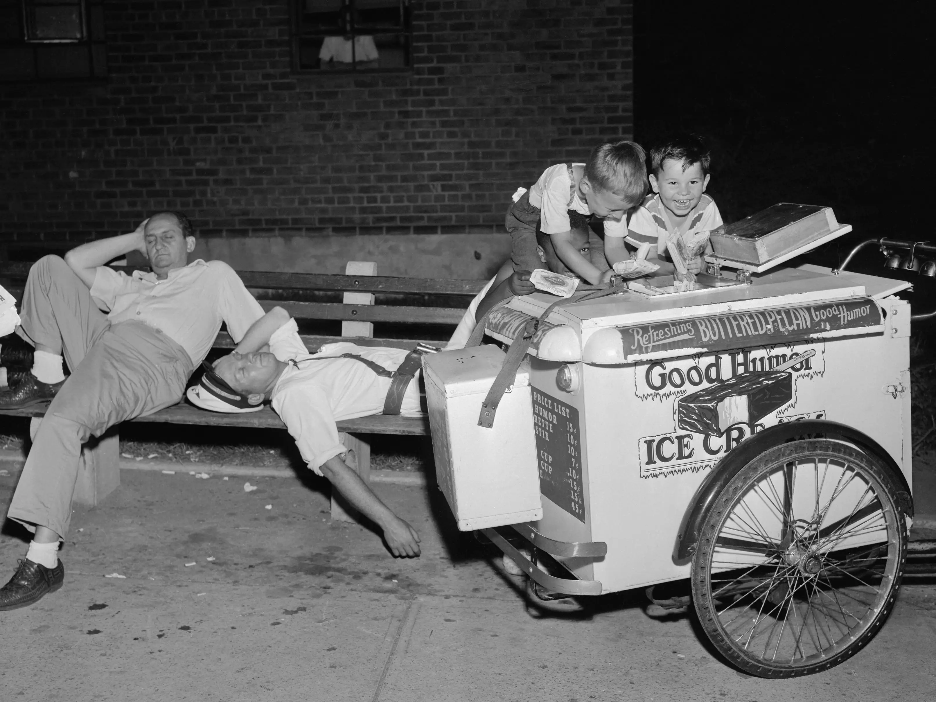 Children take ice cream from a cart while the vendor rests on a bench.