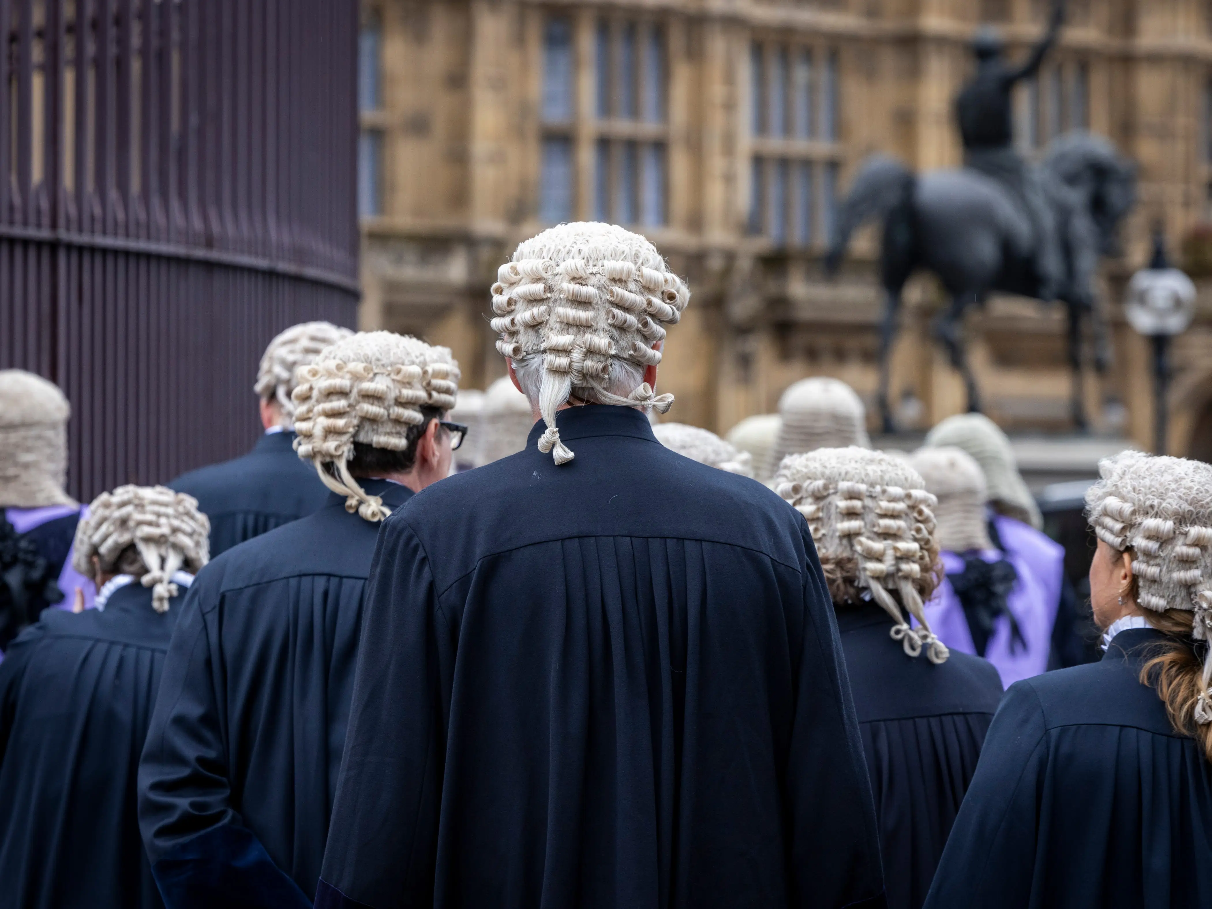 Judges in white wigs and black robes walk past a historic government building.