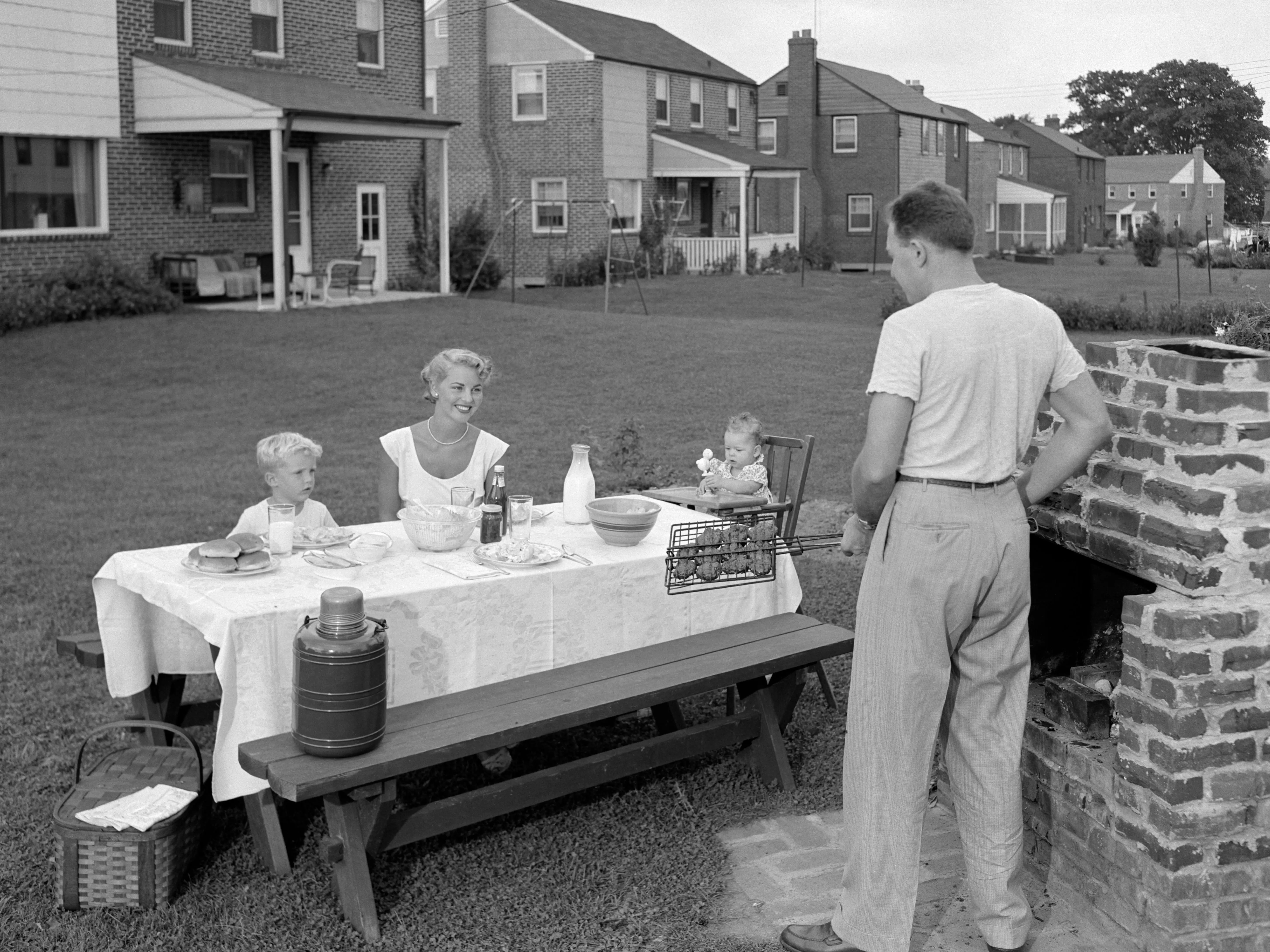 A mother and two children sit at a table while a husband stands at an outdoor stove