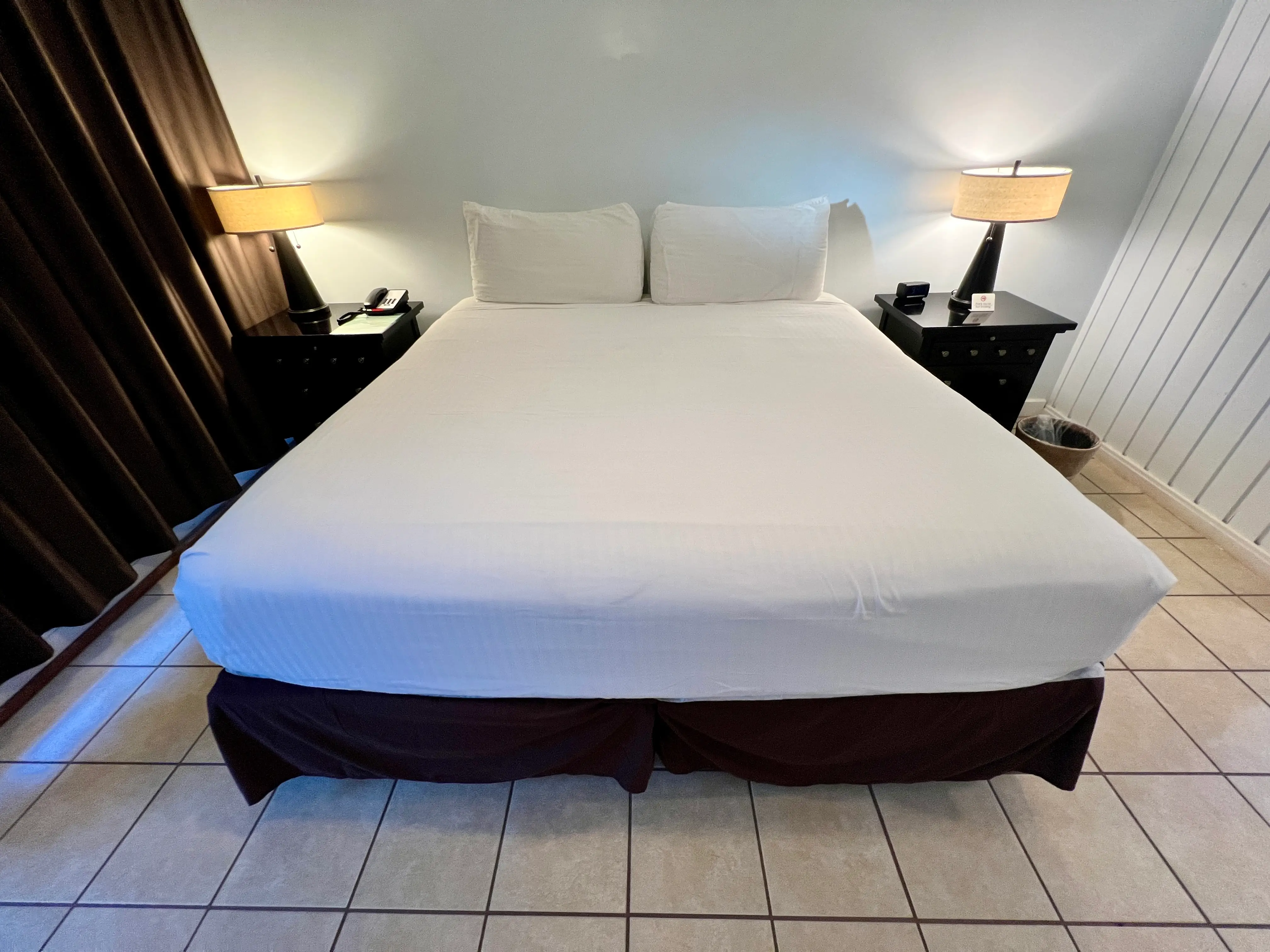View of king-size bed with white sheets, dark wood nightstands with lamps on top on either side of it