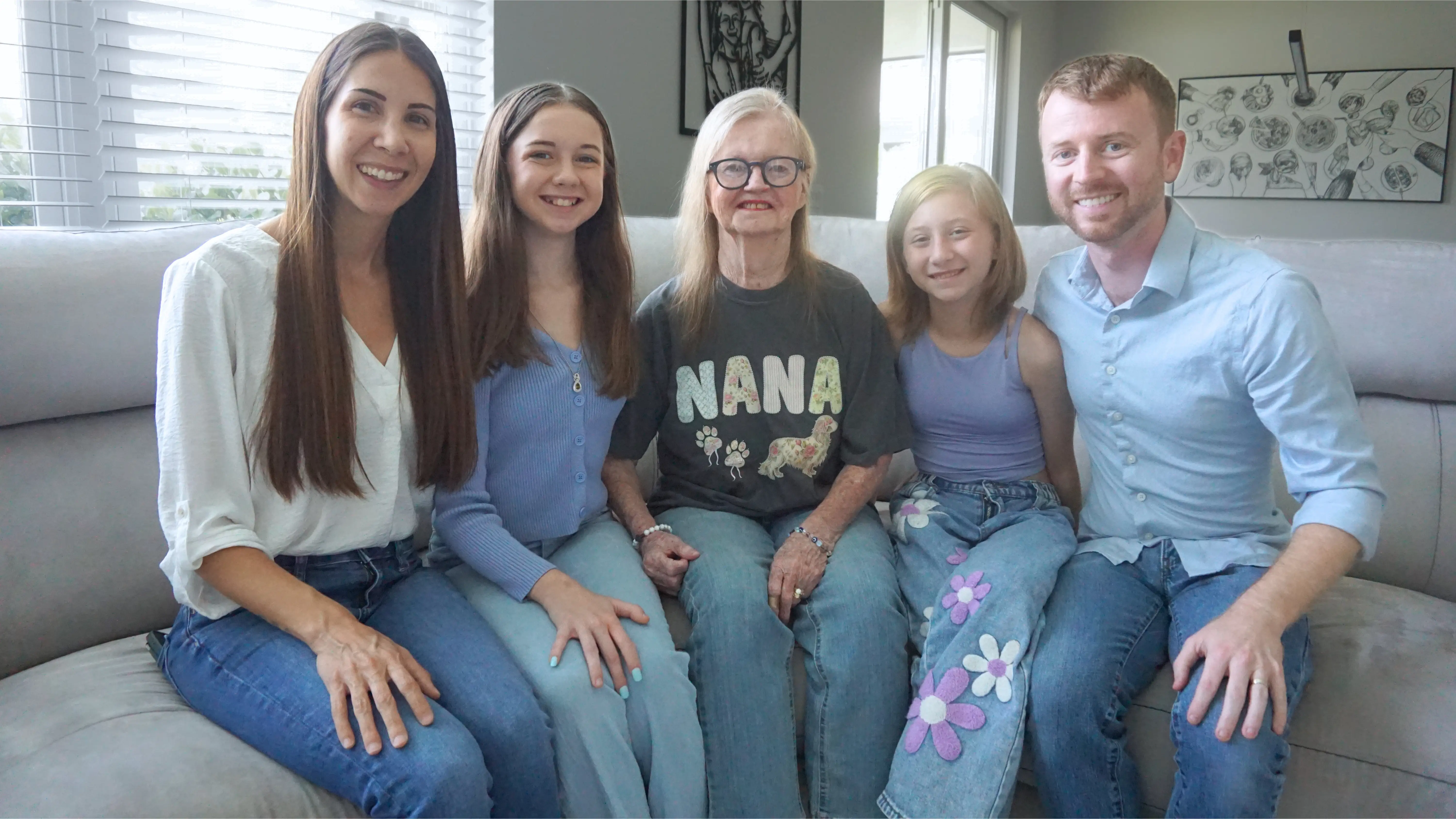 Five people sit together smiling on a light-colored couch in a living room, with one shirt reading “NANA.”