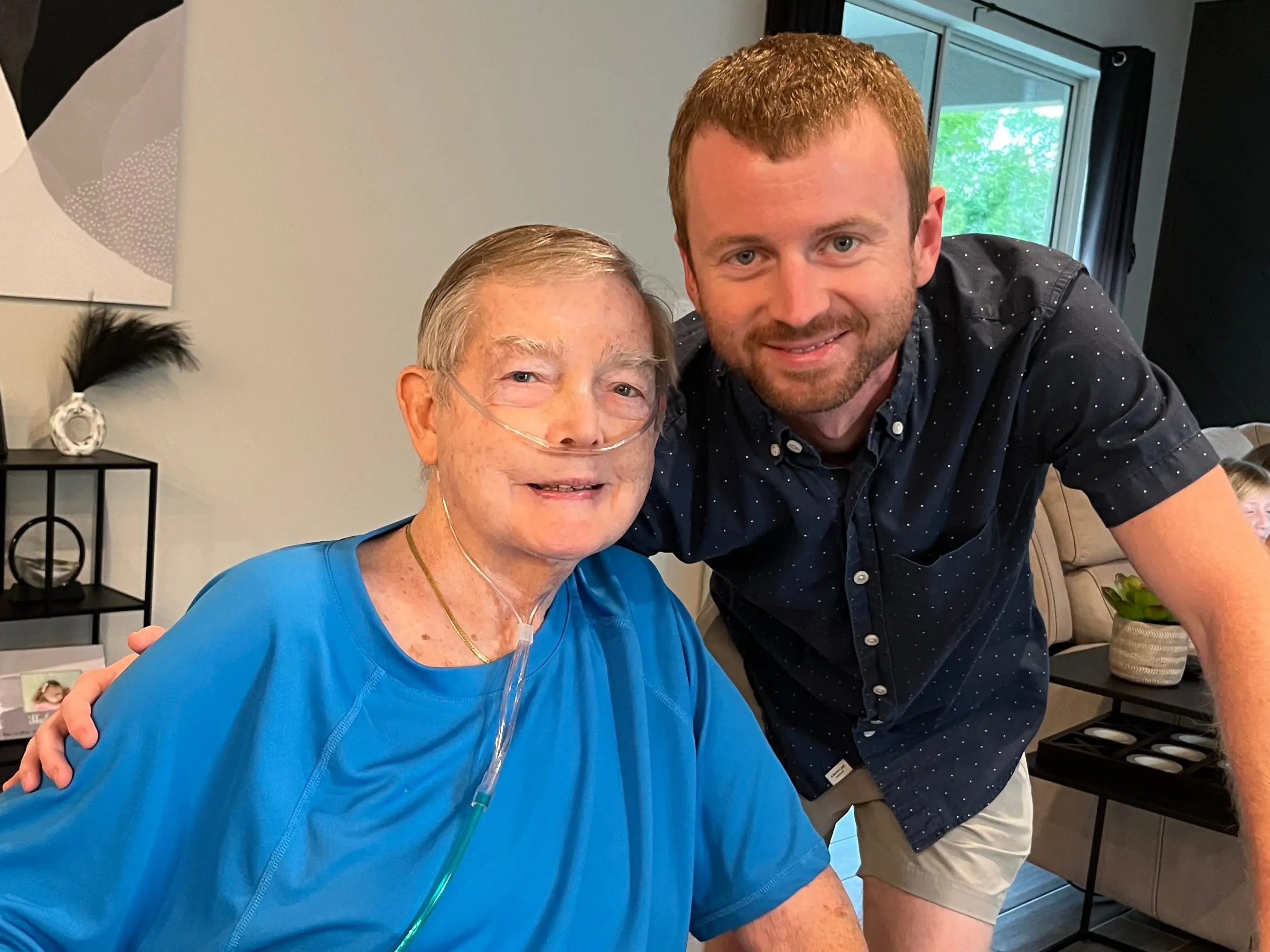 Two people pose in a living room, with one seated wearing a nasal cannula and blue shirt.