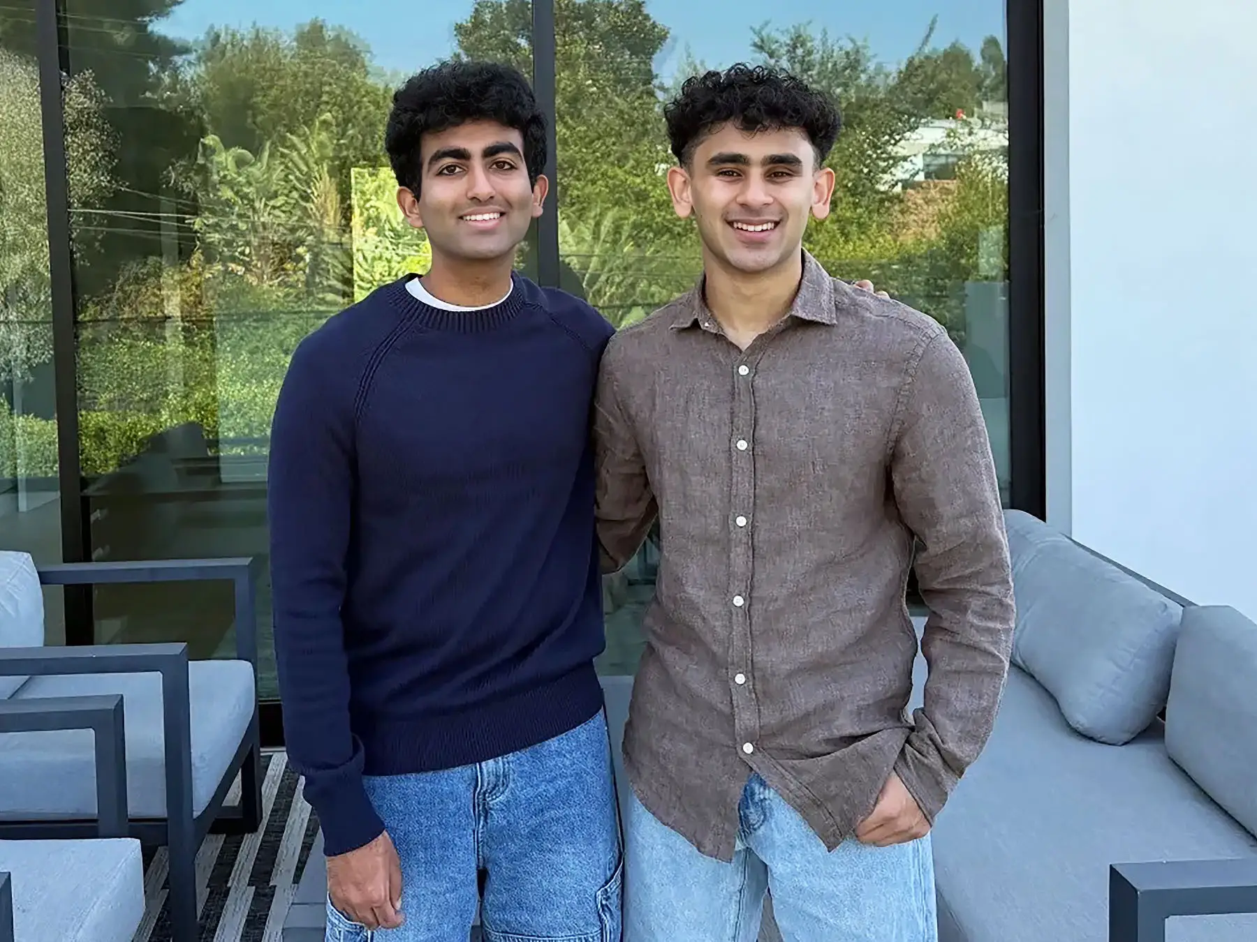 Two smiling people stand together on a patio in front of large windows and outdoor seating.
