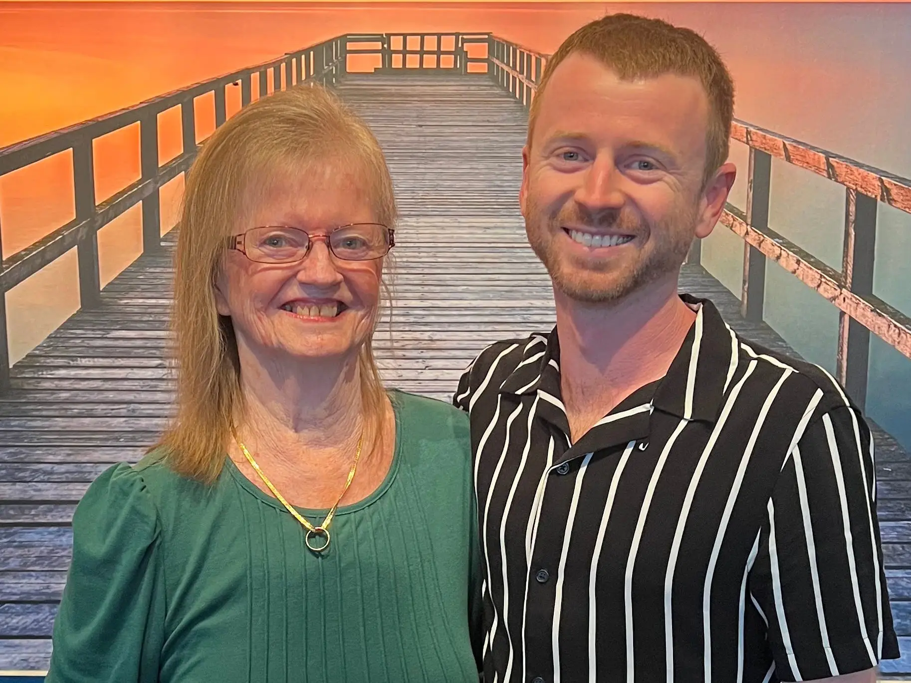 Two smiling people pose arm in arm in front of a sunset pier mural indoors.