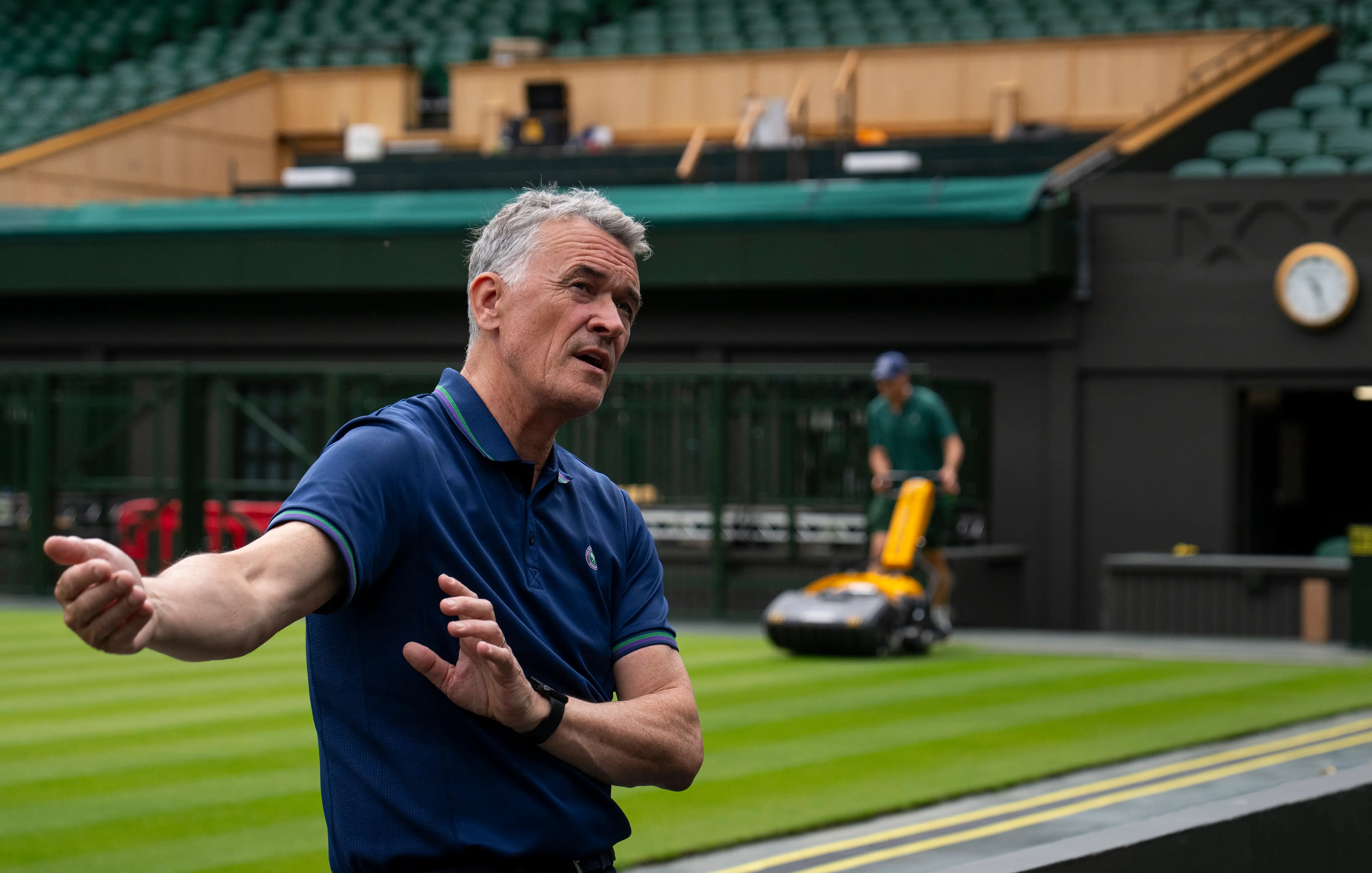Neil Stubley, Head of Courts and Horticulture at Wimbledon, stands in front of Centre Court while a groundskeeper mows the lawn in the background.