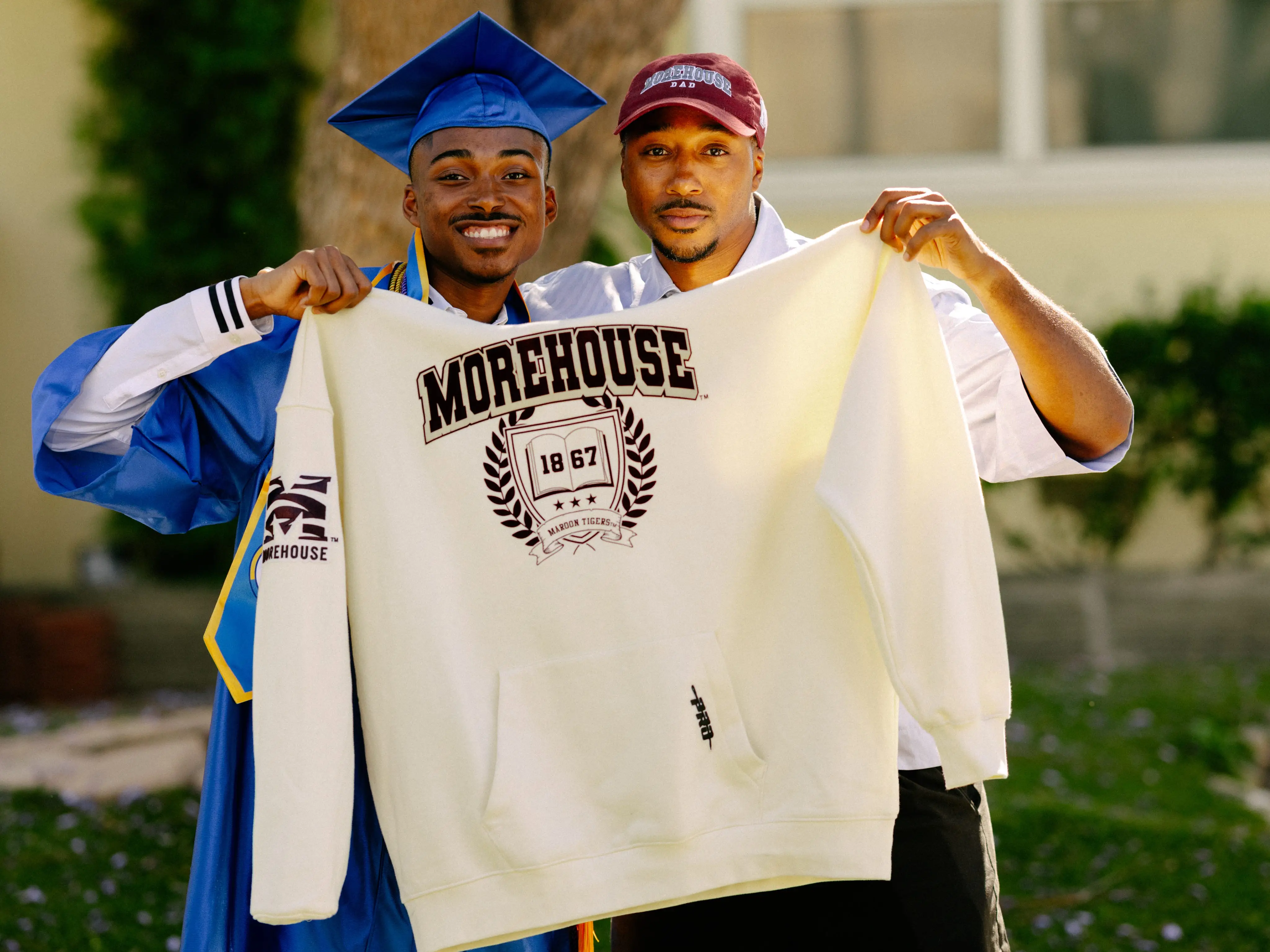 Two people hold a MOREHOUSE sweatshirt outdoors beside a graduate in a blue cap and gown.