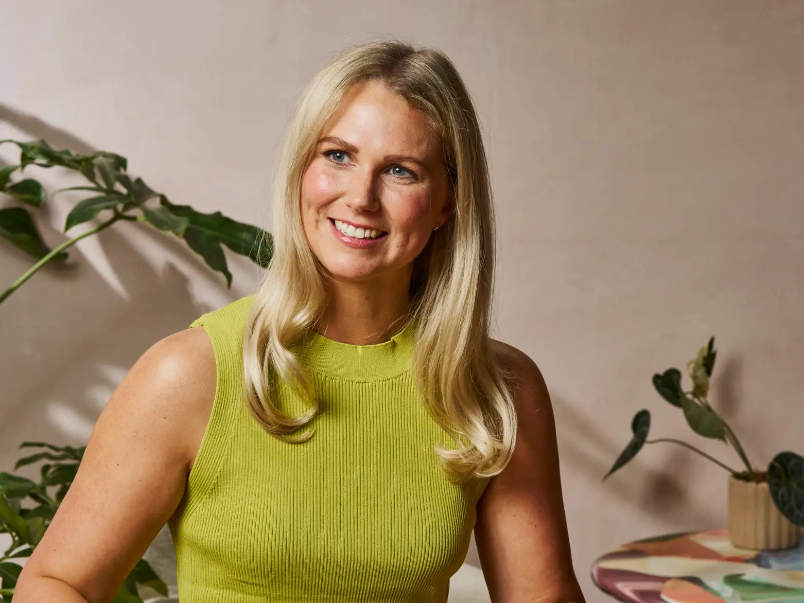 Smiling person in a lime sleeveless top serves a colorful skillet dish at a table with plates and plants.
