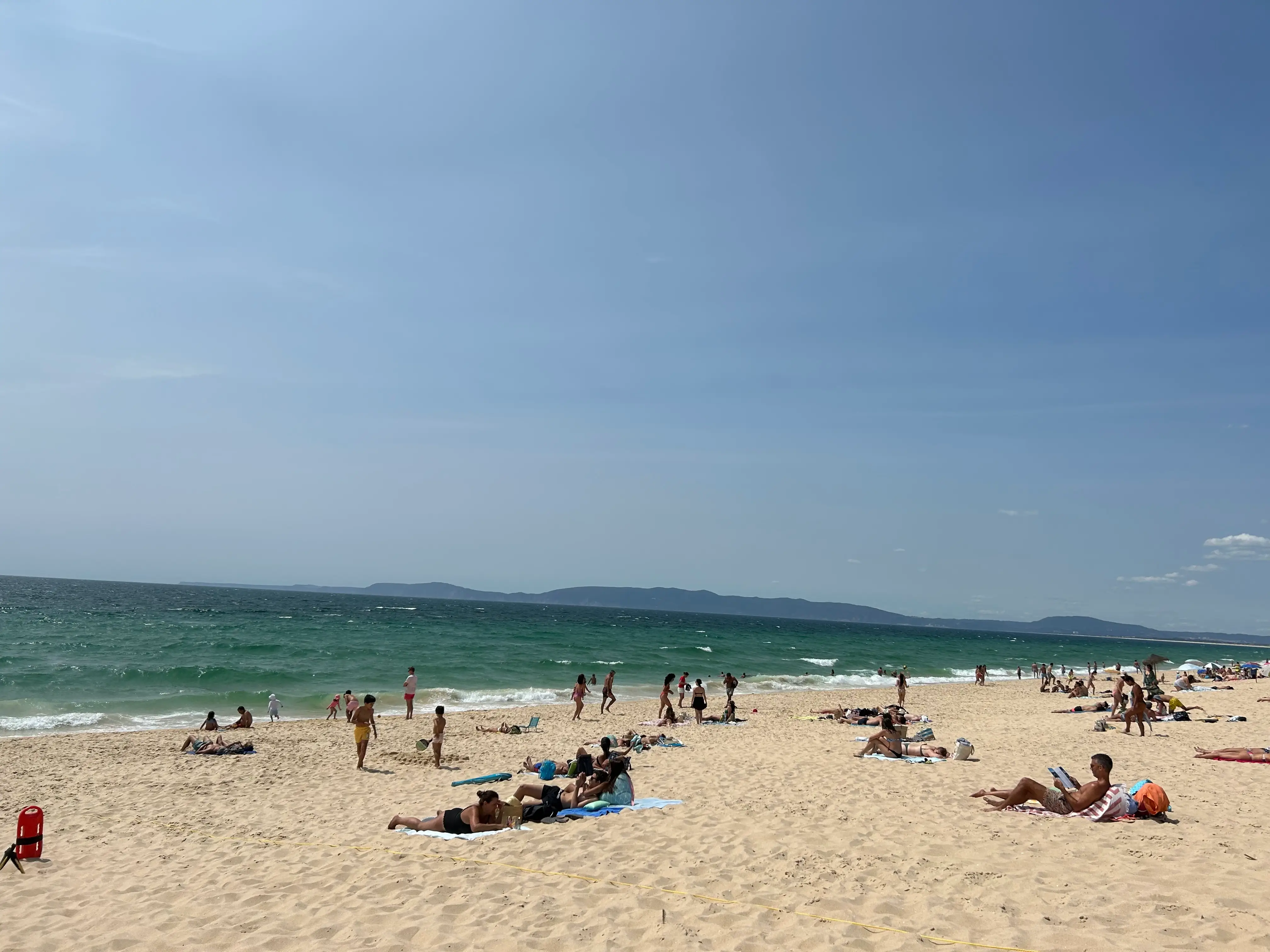 Beachgoers relax and walk on a sandy shoreline beside green-blue ocean waves under a clear sky in Comporta, Portugal.