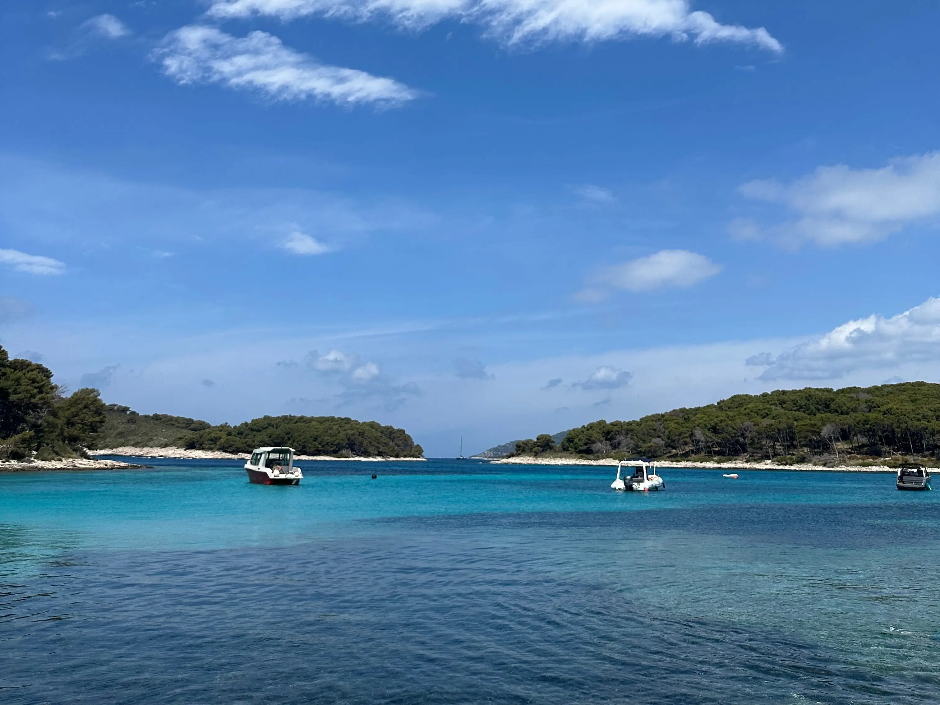 Small boats float in turquoise coastal water between tree-covered islands under a bright blue sky with scattered clouds.