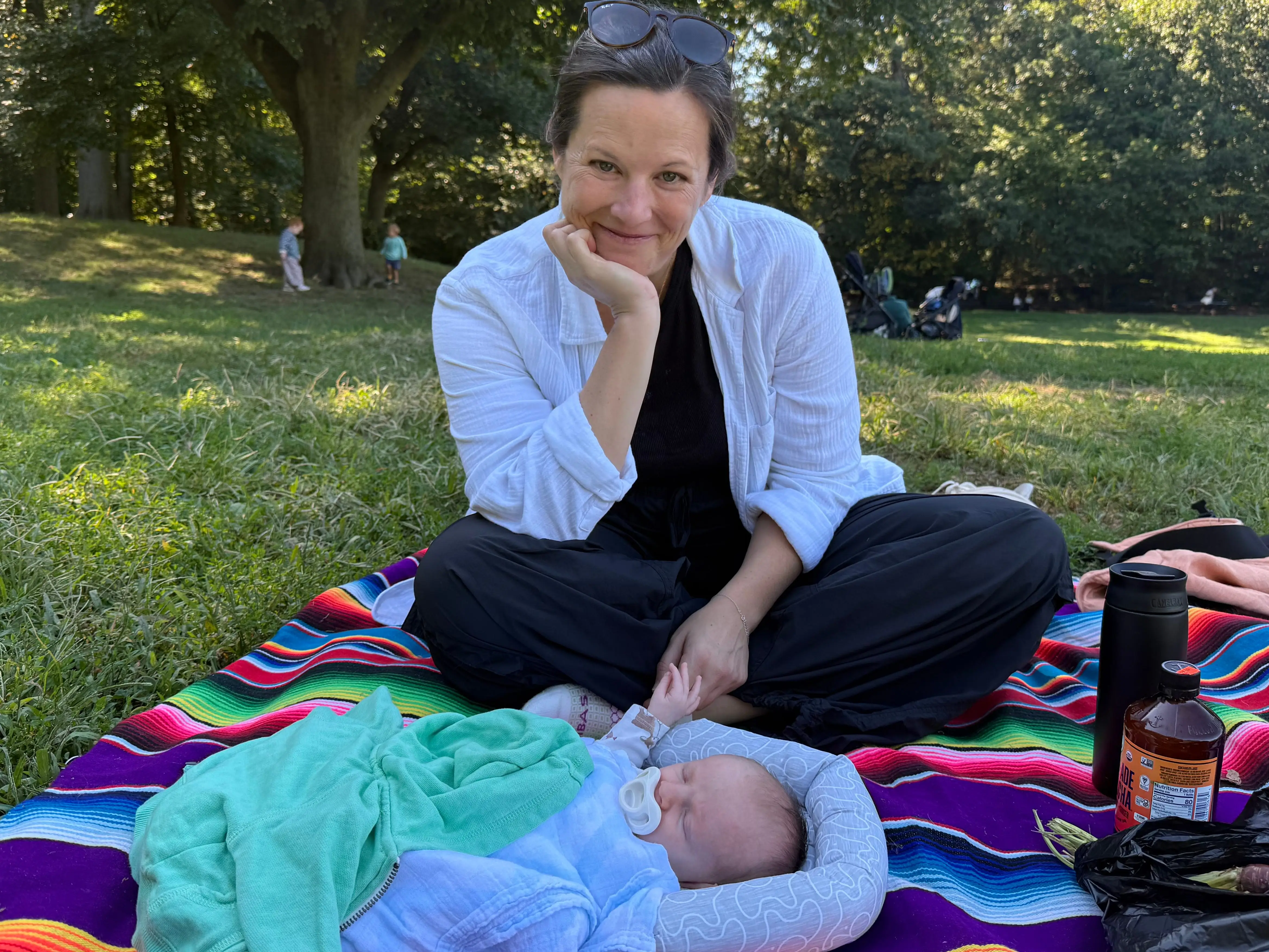 A person sits on a colorful striped blanket beside a baby in a sunny park.