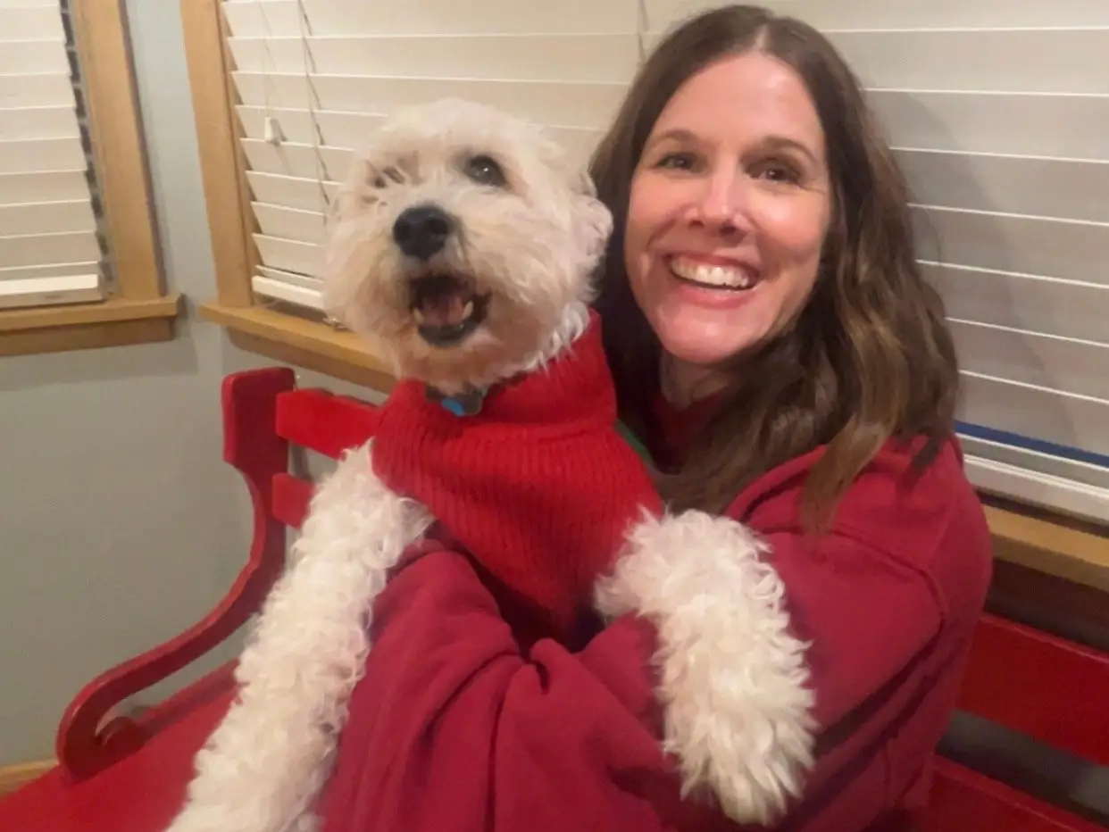 Smiling person in a red sweater hugs a white dog wearing a red sweater on an indoor bench.