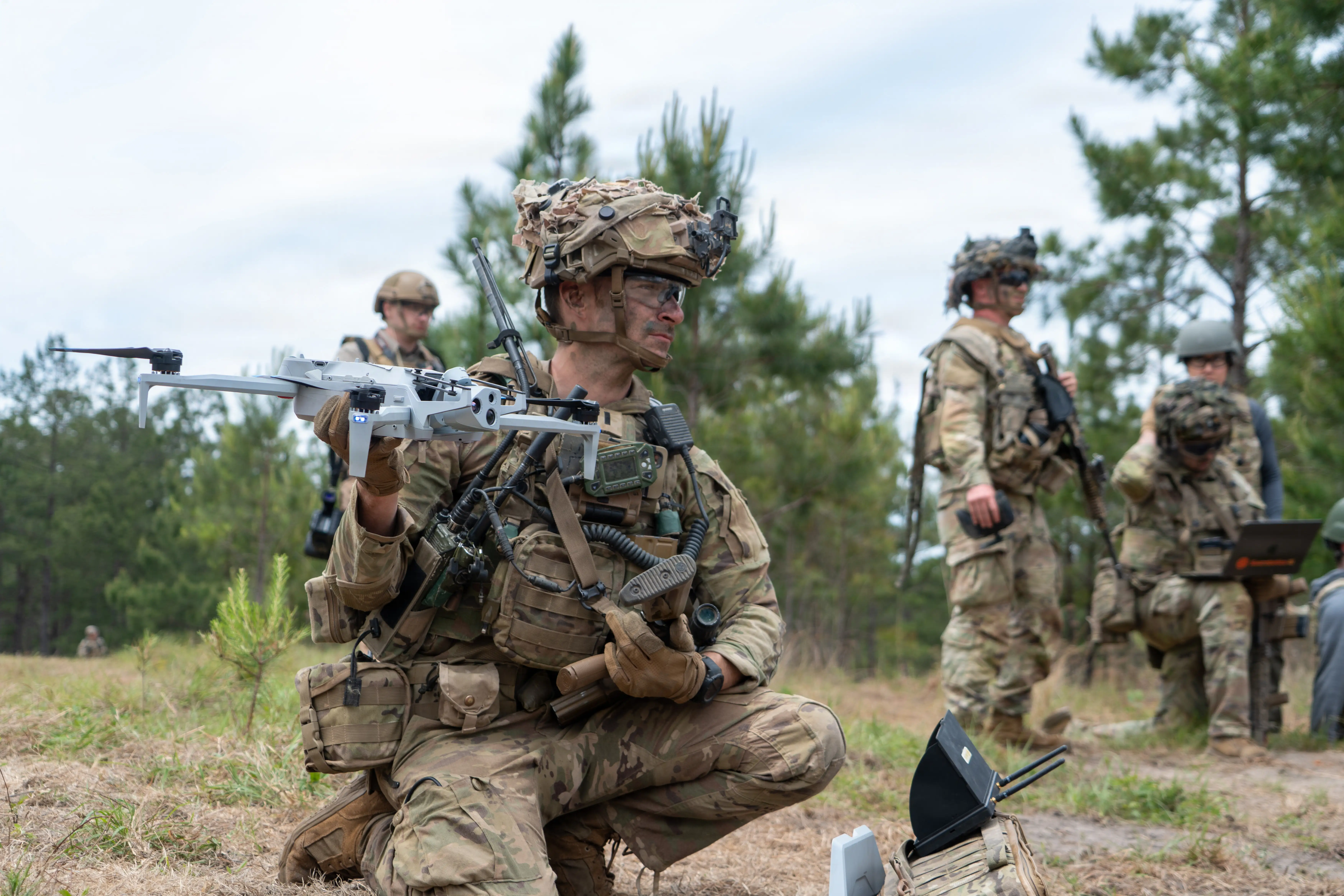 A soldier wearing camouflage holds a white drone. Three soldiers stand behind him, looking towards the right. The background is full of trees.