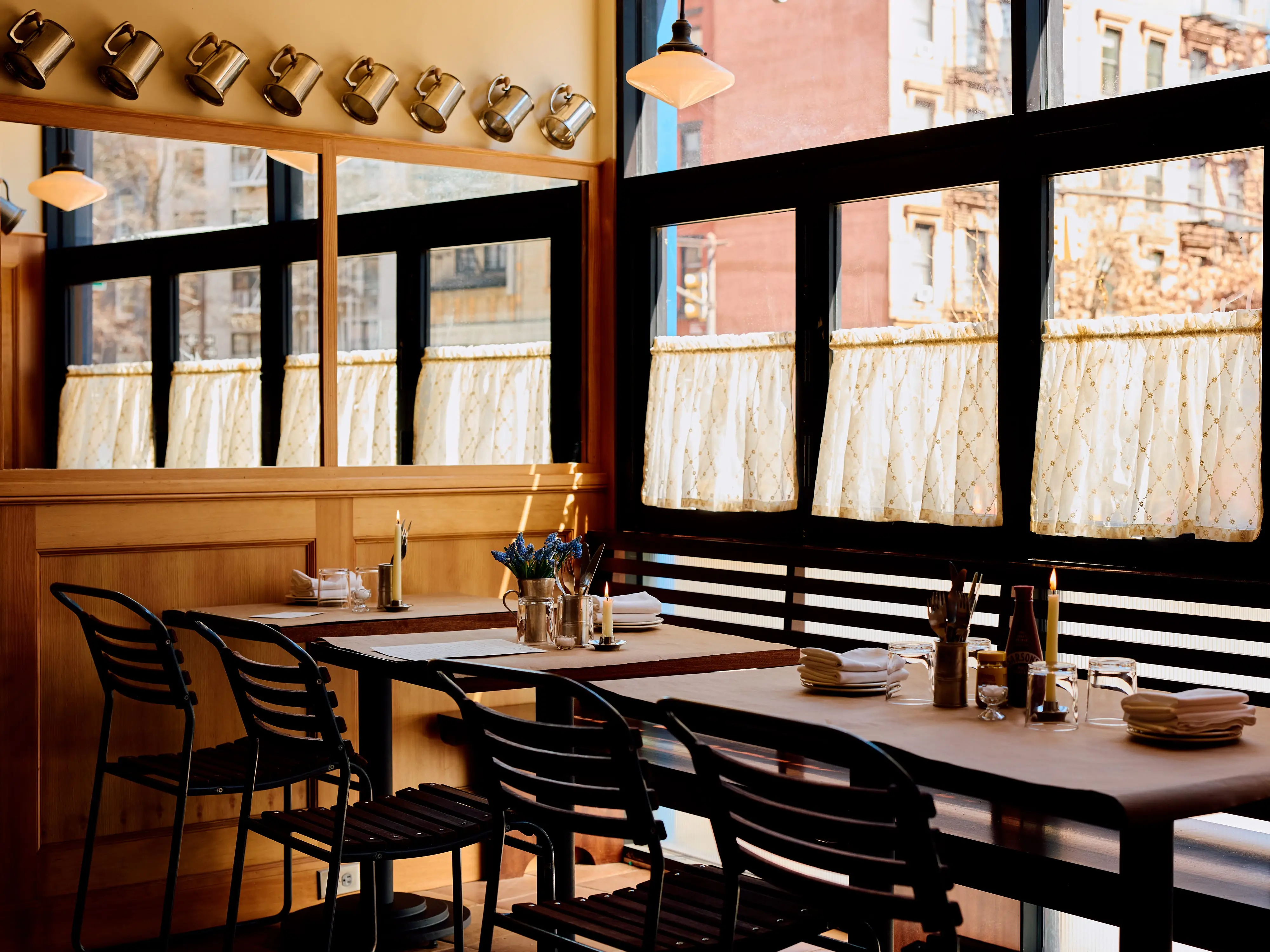 Sunlit restaurant dining area with set tables, black chairs, wood paneling, mirrors, and lace-curtained windows at Dean's.