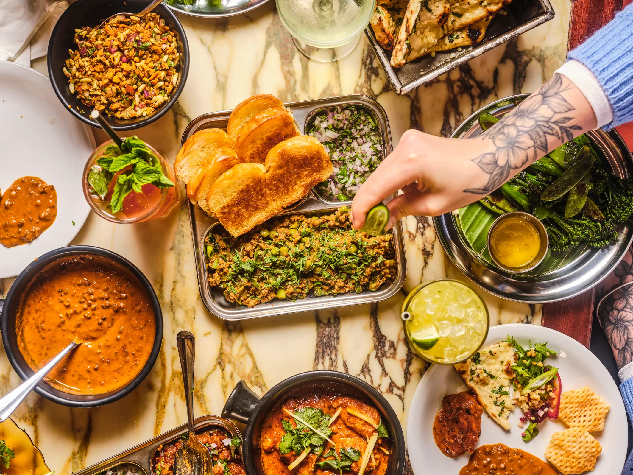 Overhead table spread of assorted curries, breads, vegetables, drinks, and a tattooed hand squeezing lime.