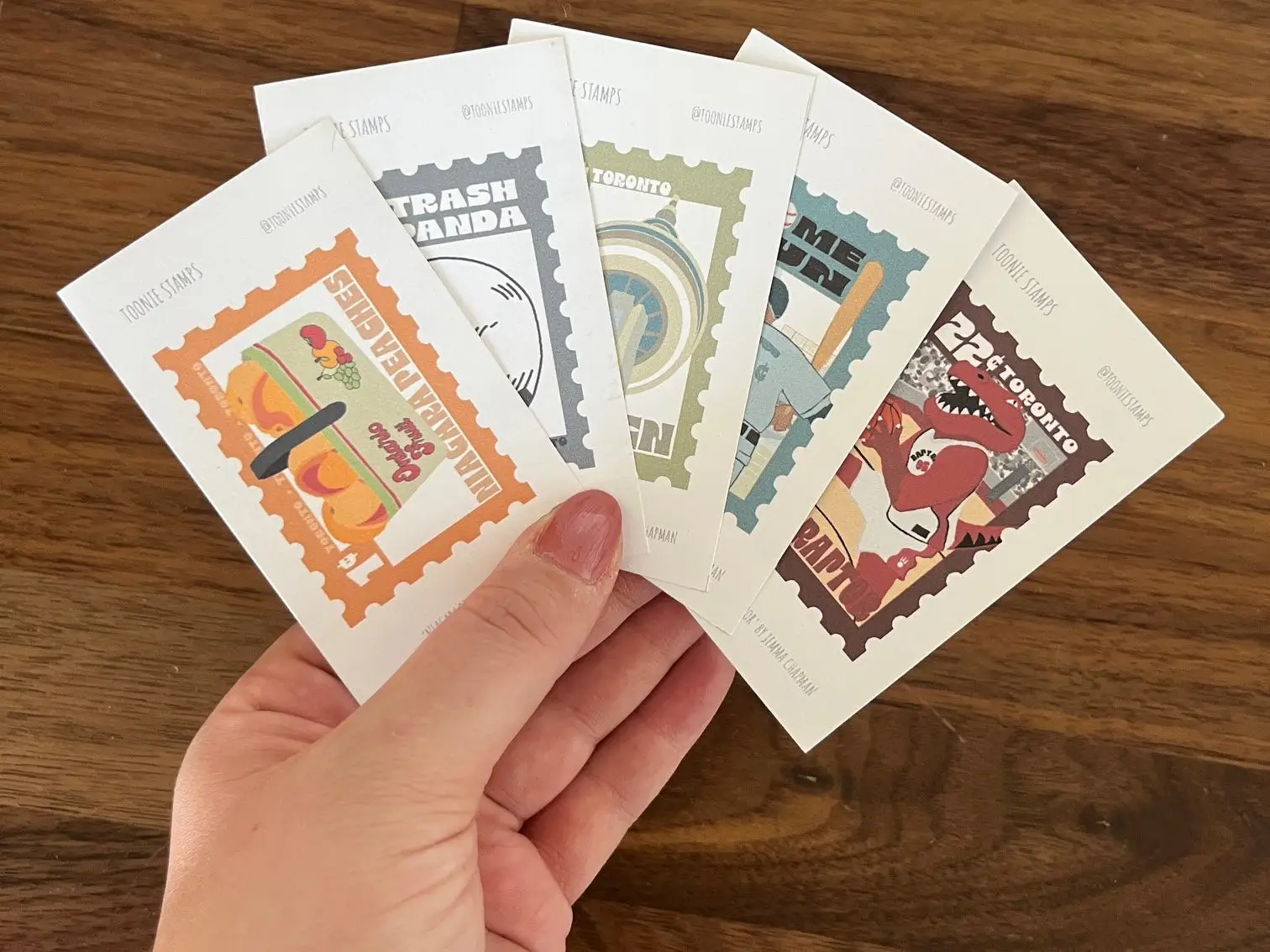A hand fans out illustrated stamp-style cards over a wooden floor.