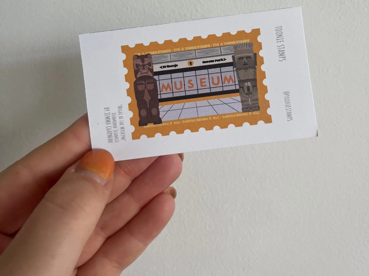 A hand holds a white card with a stamp-style museum illustration against a plain wall.