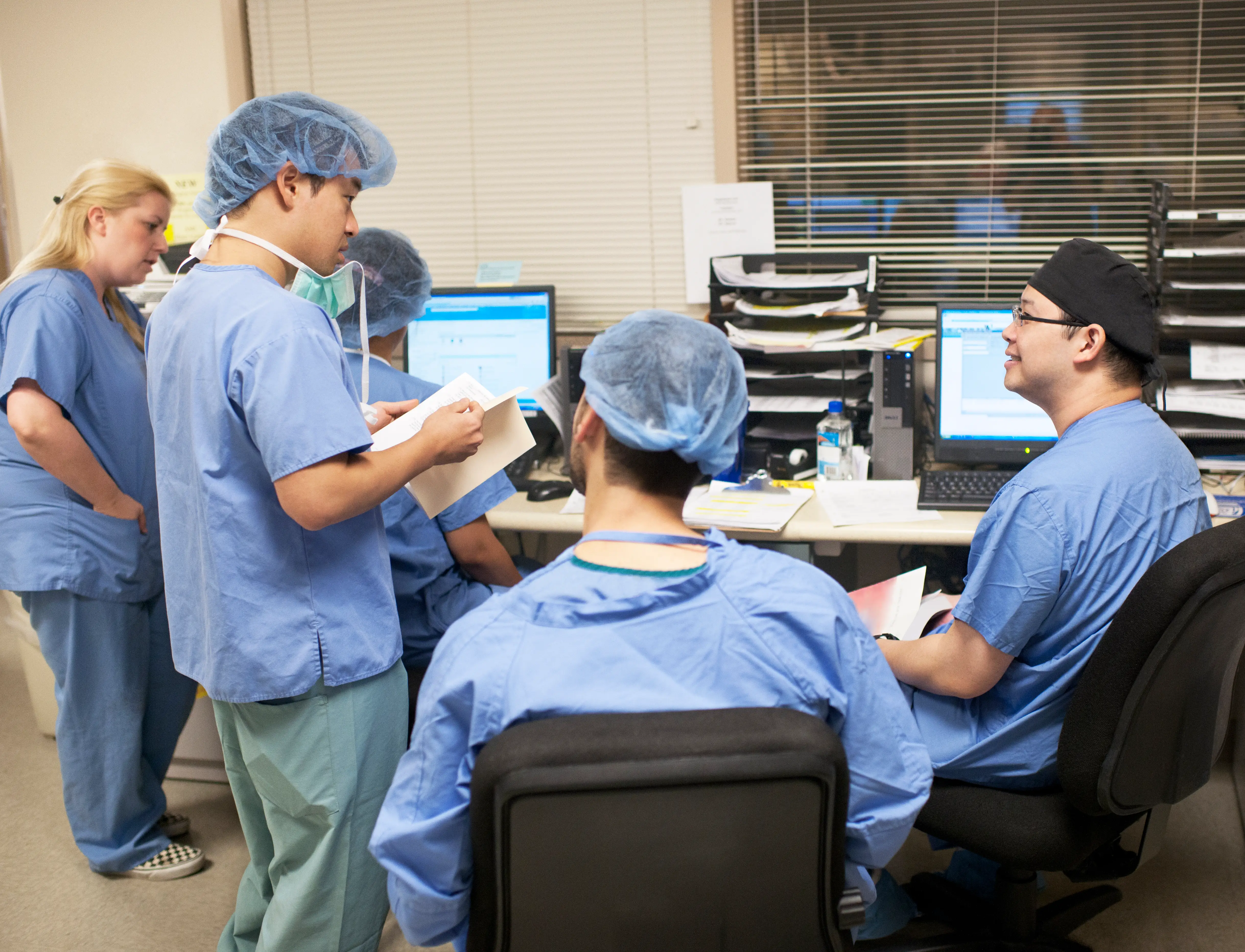 Healthcare staff in blue scrubs, and most with hair covers, gather around computers