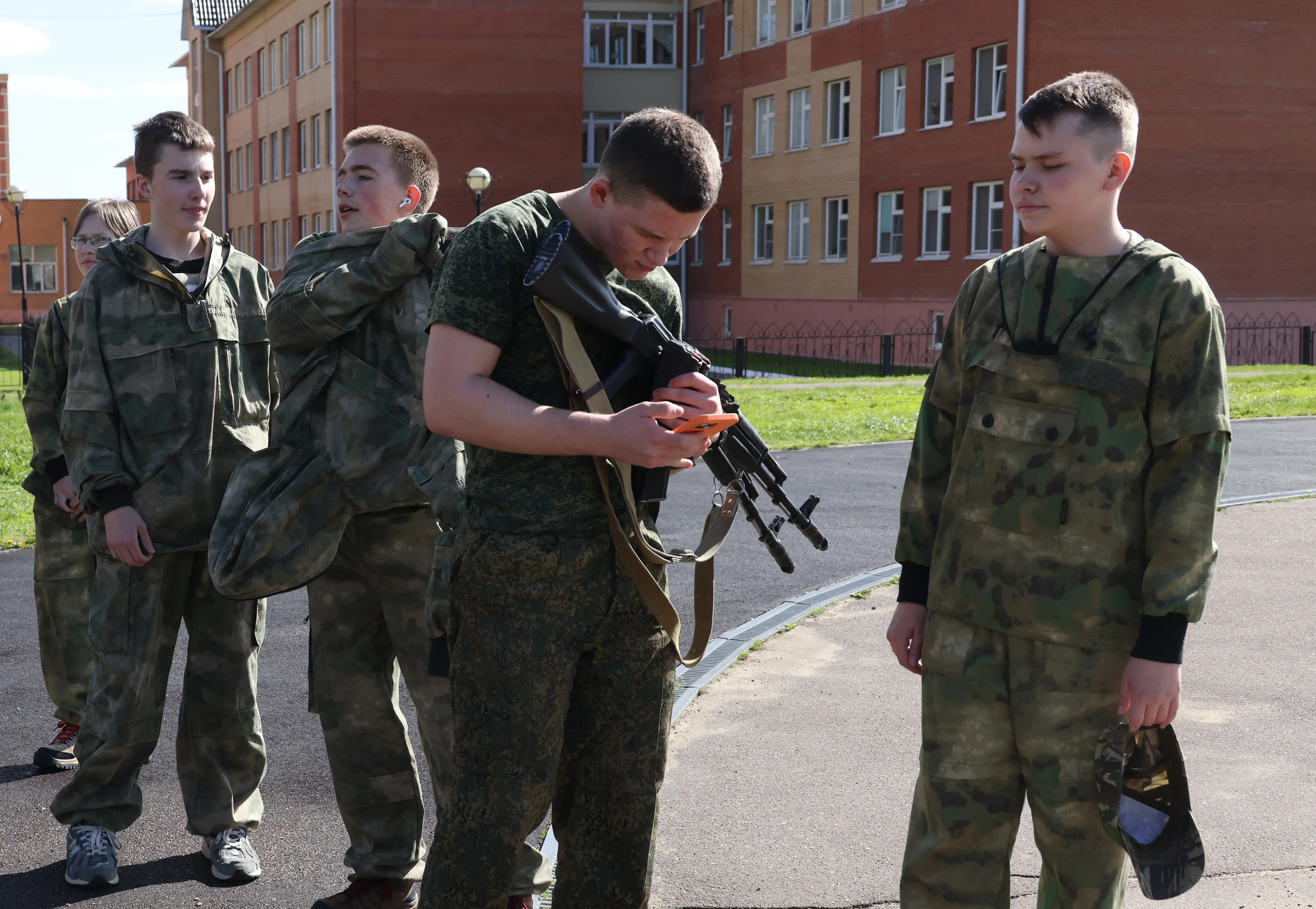 A Russian teenager looks at his phone while holding a Kalashnikov rifle.