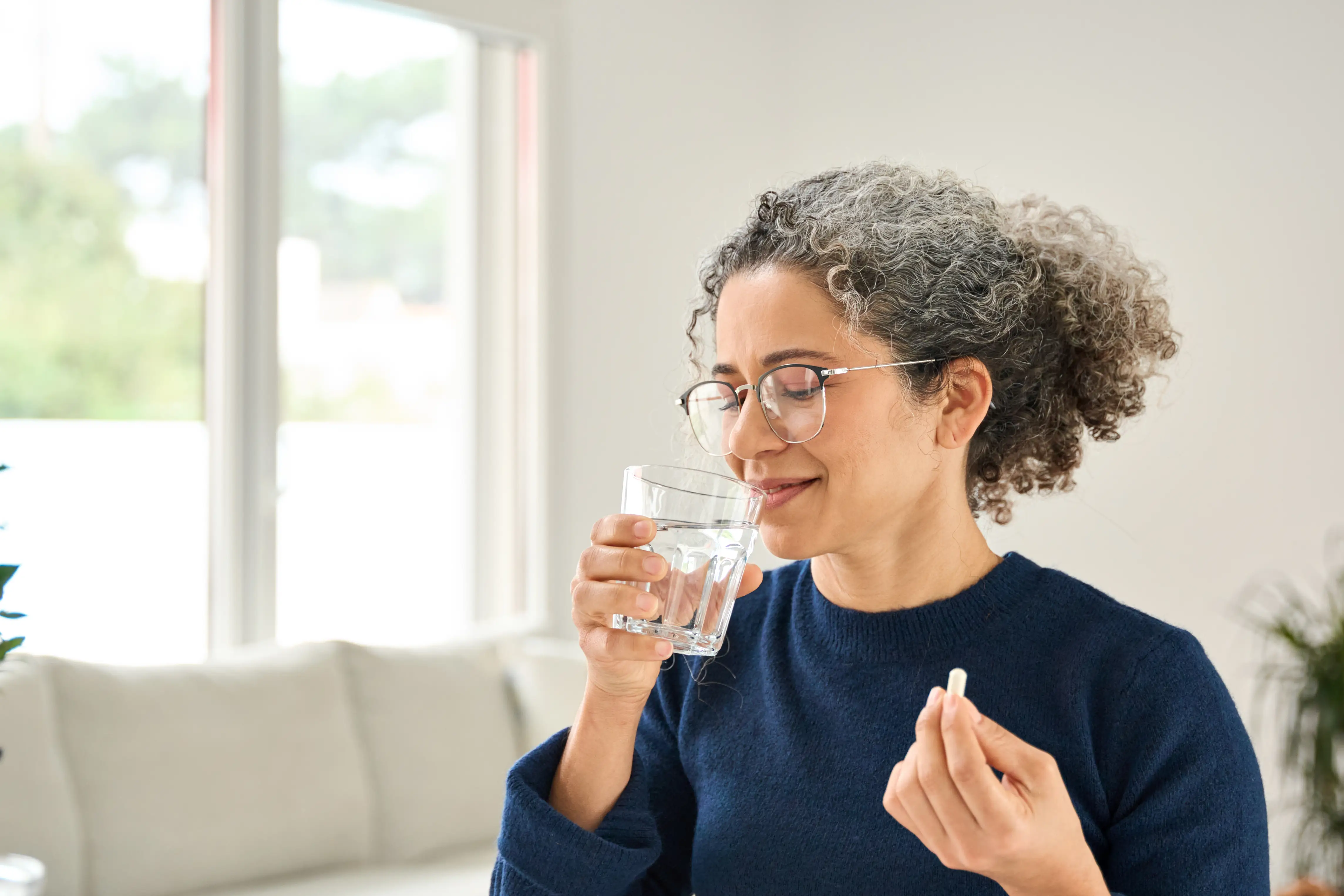 A woman drinks from a glass of water while holding a pill in one hand.