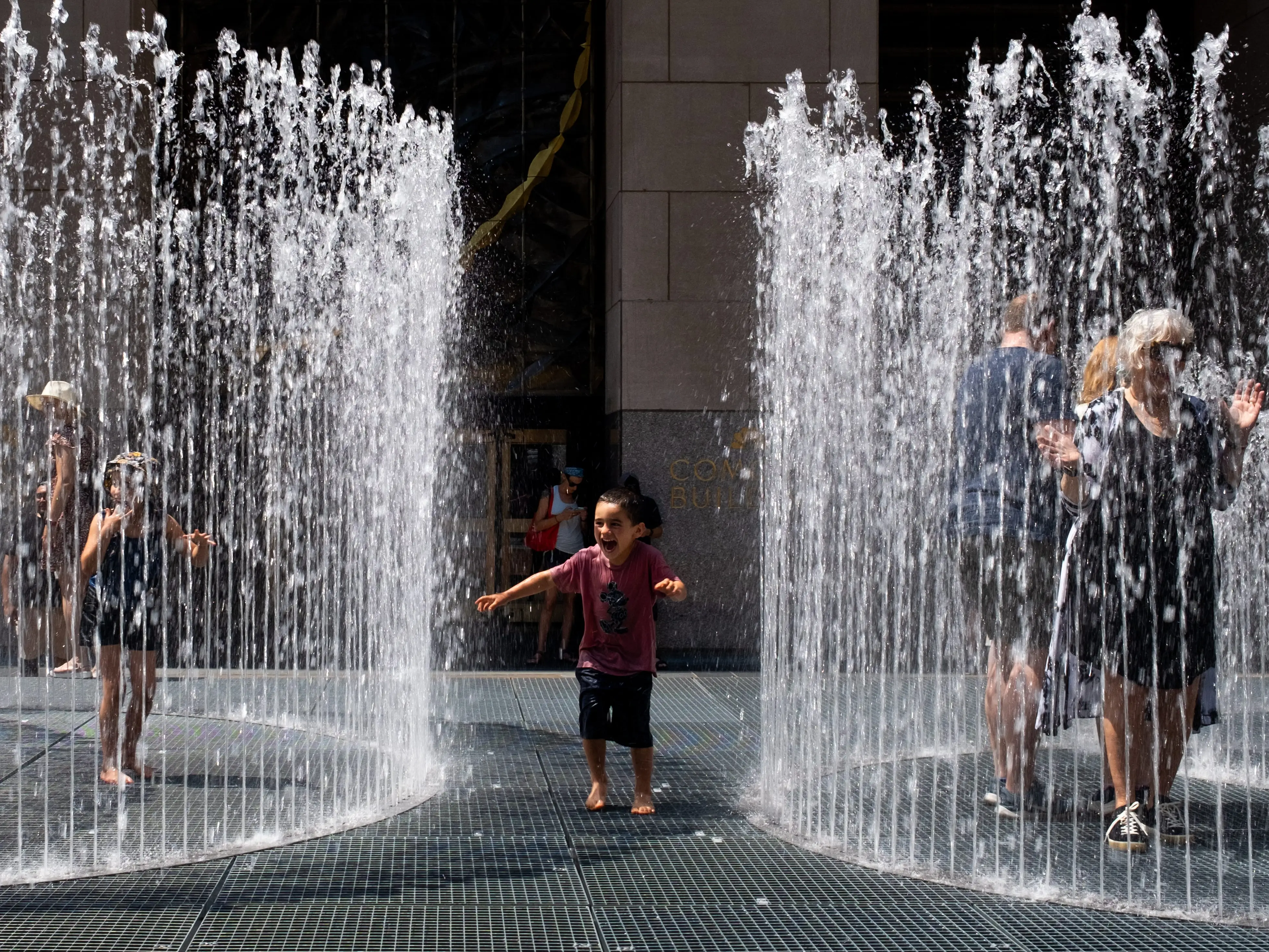 Several people play and walk among tall outdoor fountain jets on a sunny paved plaza.
