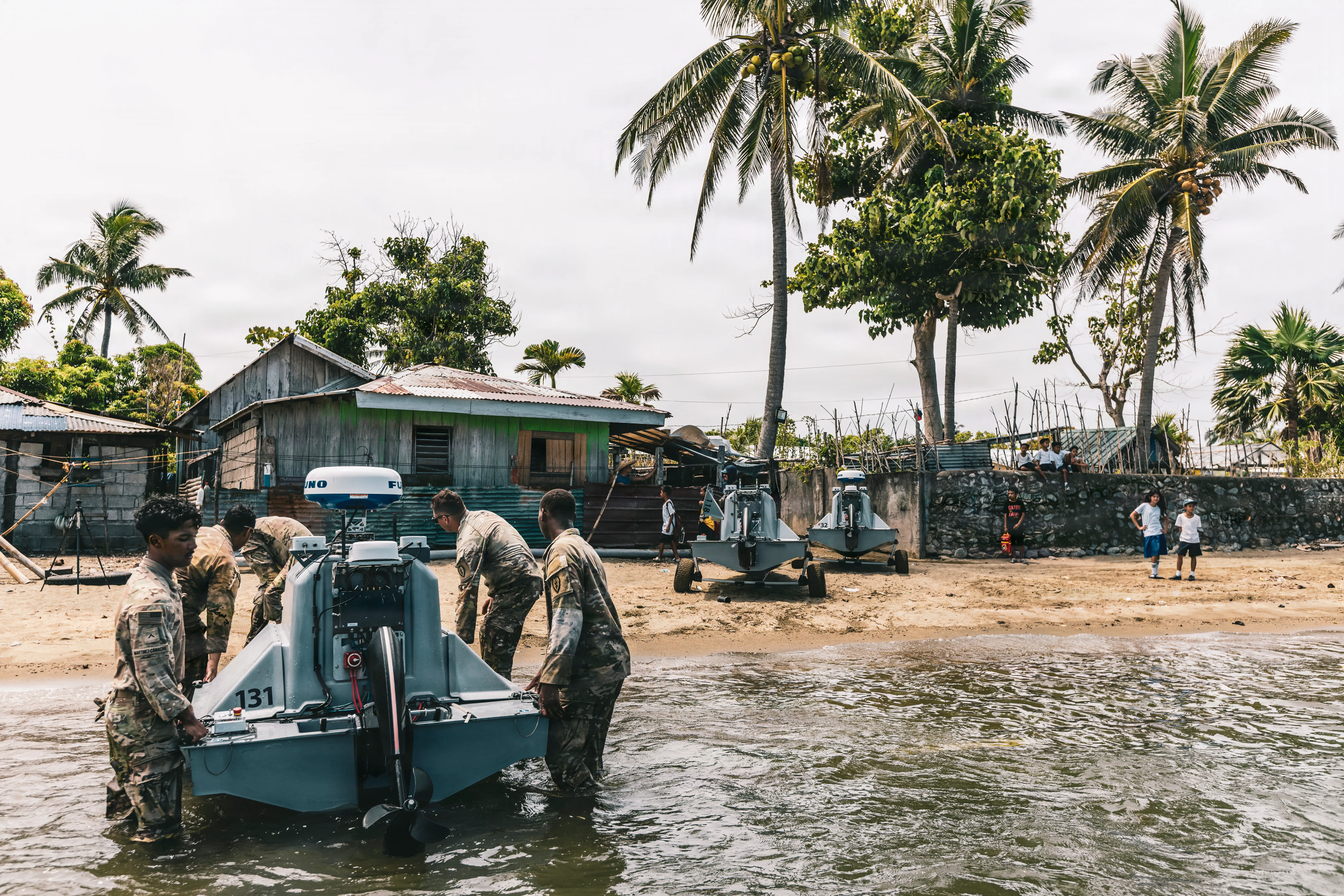 US Army soldiers carry a water drone onto a beach in the Philippines with houses and trees.