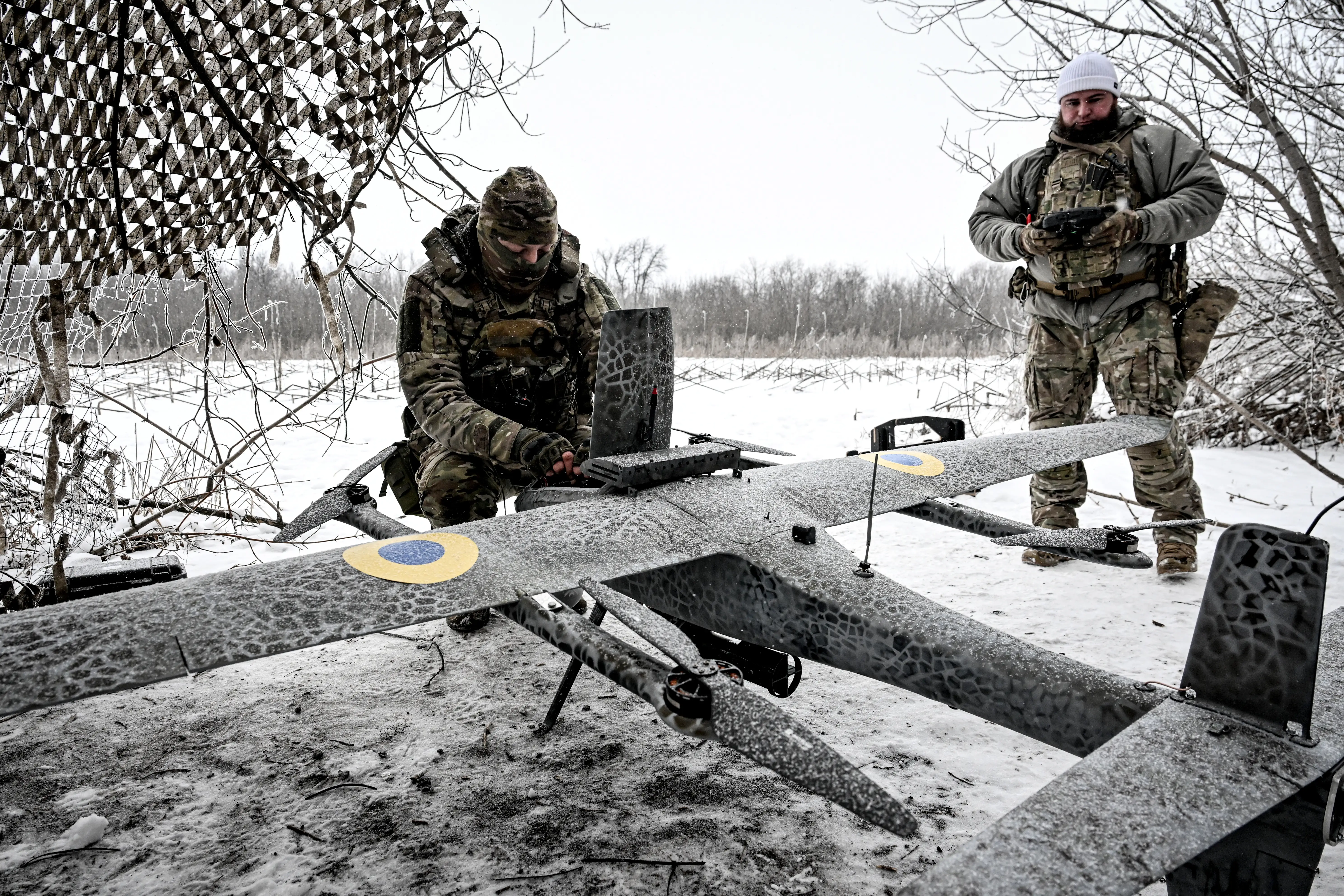 Two men in camouflage gear in a snowy field under a net beside a large drone
