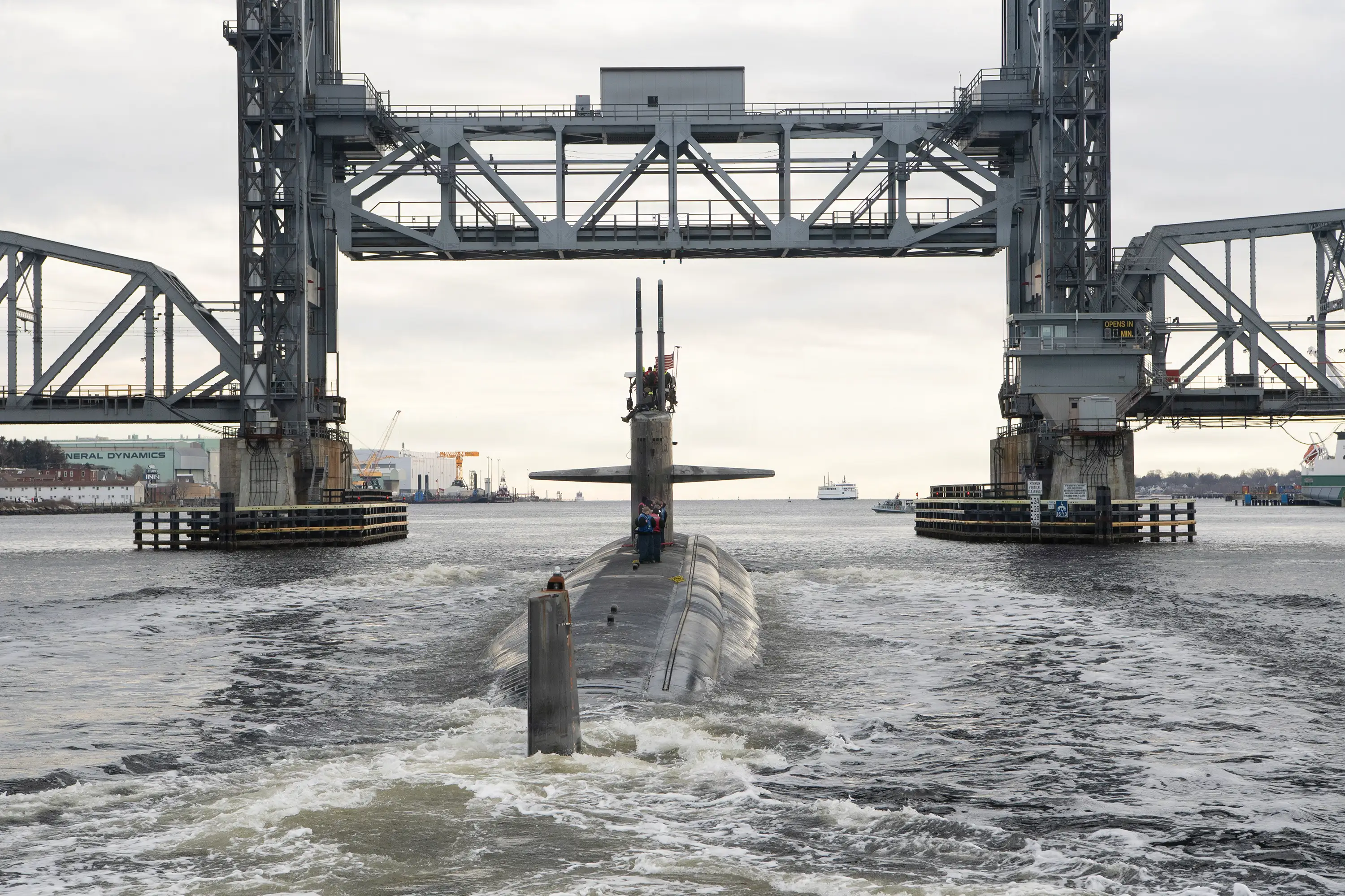 A submarine sails in dark blue water under a bridge.