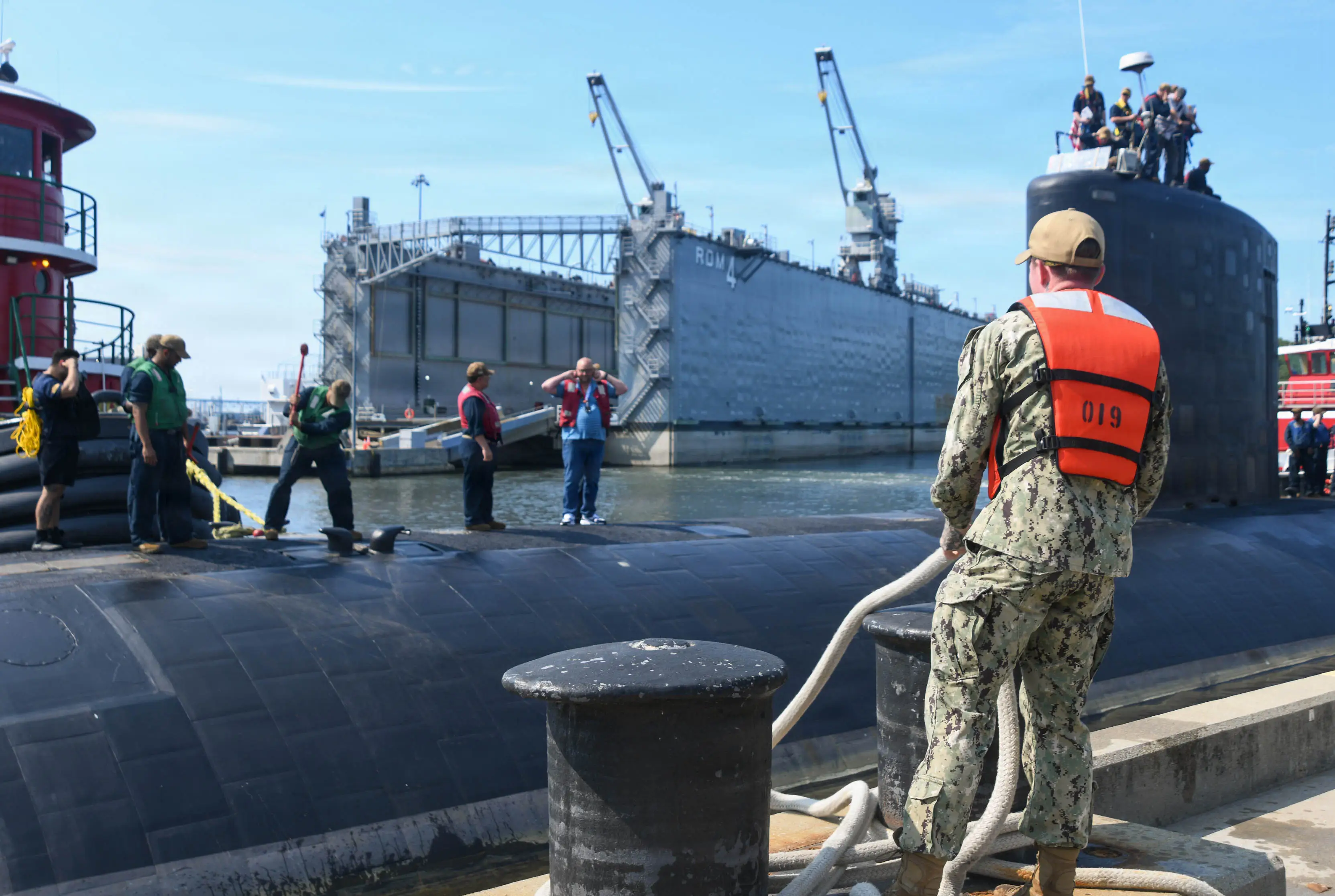 A group of men stand on top of and around a black submarine docked.
