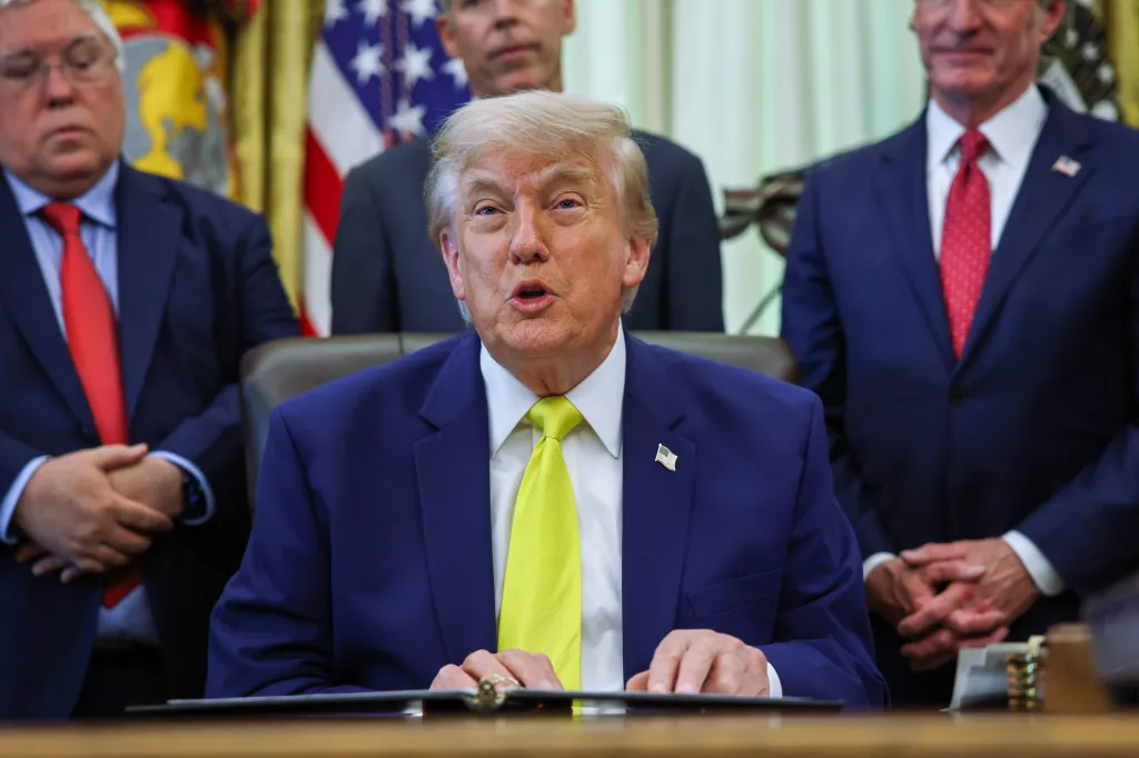 Donald Trump speaking from behind a desk while two men stand behind him.
