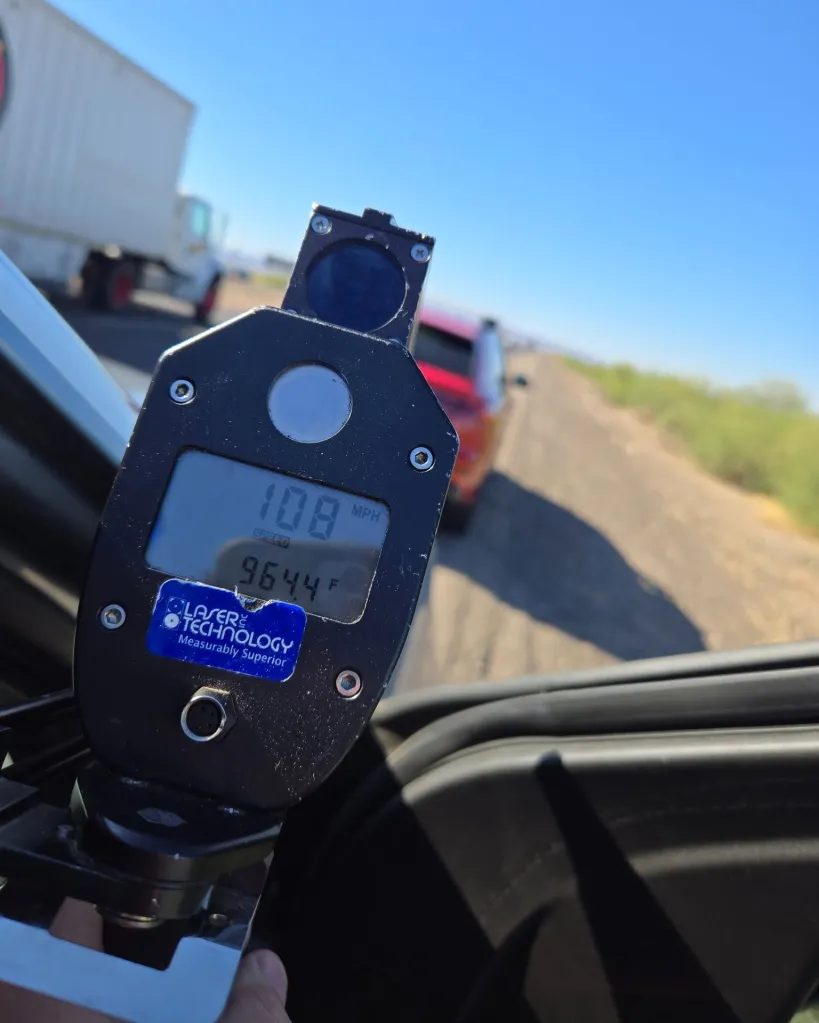 A radar gun displaying a speed of 108 MPH and a temperature of 96.4 F, viewed from inside a vehicle on a sunny day with another vehicle in the distance.
