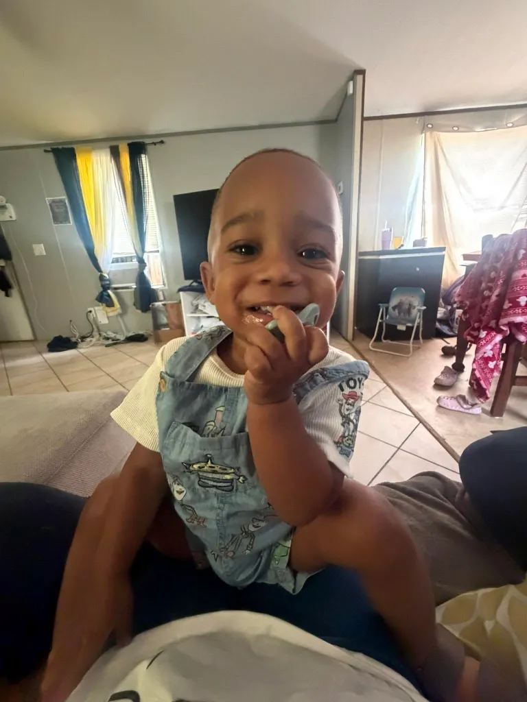 A young child in overalls smiles at the camera while chewing on a blue toy.