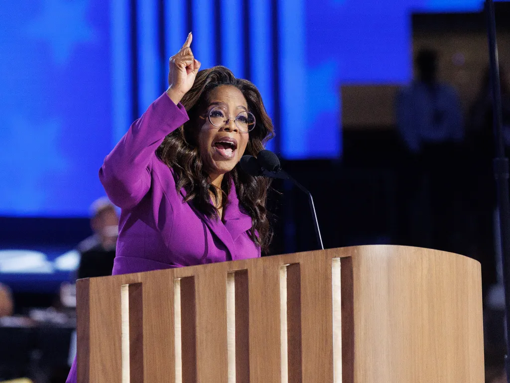 Oprah Winfrey speaking at a podium with her arm raised and mouth open.