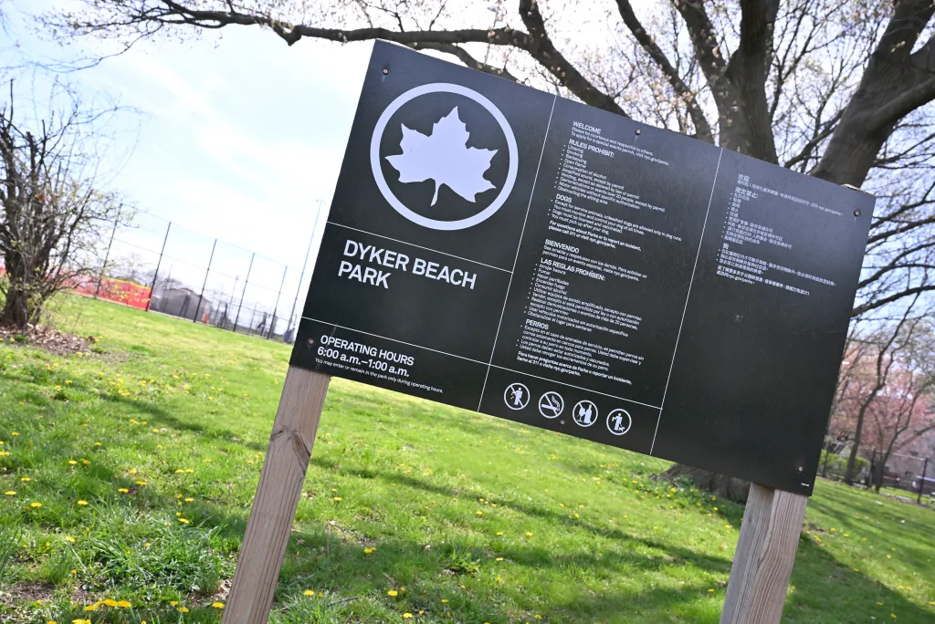 A sign for Dyker Beach Park listing rules in multiple languages, with a grassy field and trees in the background.