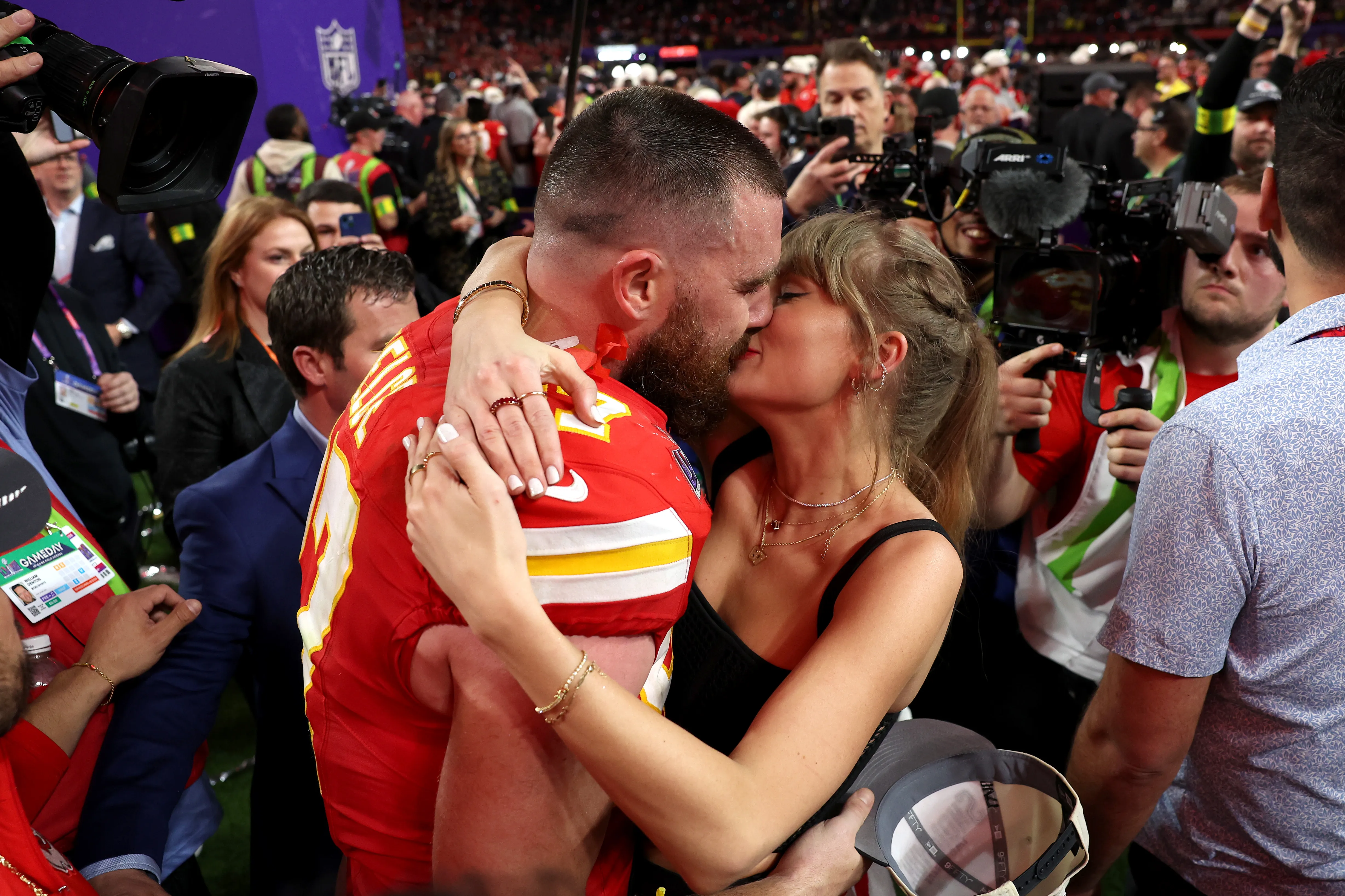 Travis Kelce and Taylor Swift embracing and kissing on a football field.