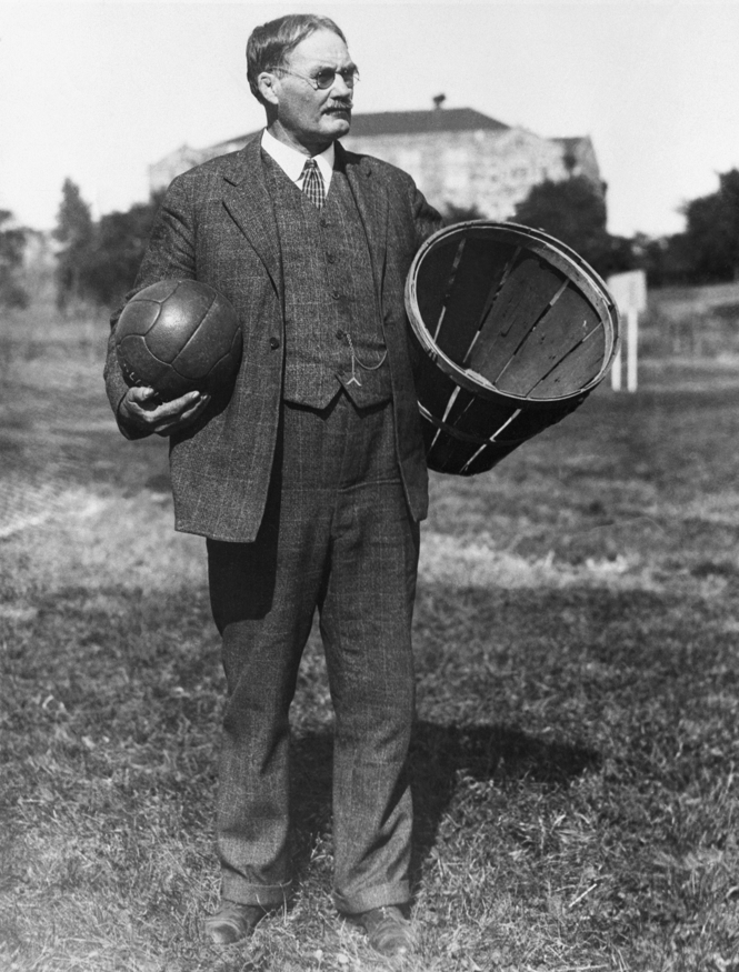 archival black-and-white photo of man standing outside wearing glasses and 3-piece tweed suit and tie holding a ball in one hand and a peach basket in the other