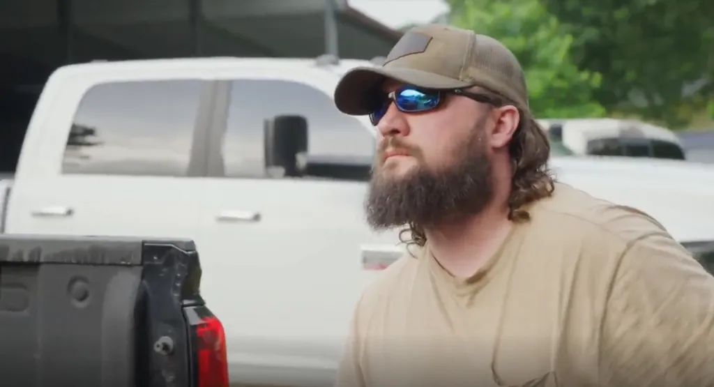 Man in a baseball cap and sunglasses with a beard and mullet, standing in front of a white truck.
