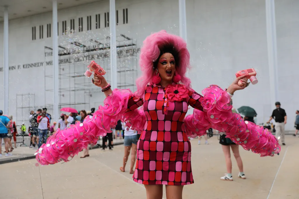 A drag queen in a pink wig and colorful dress blows bubbles from two bubble guns outside the Kennedy Center.