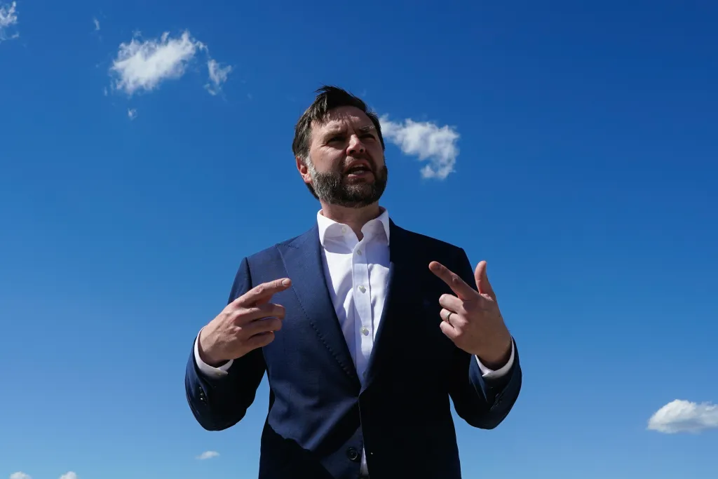 J.D. Vance in a suit, gesturing with his hands against a clear blue sky with a few clouds.