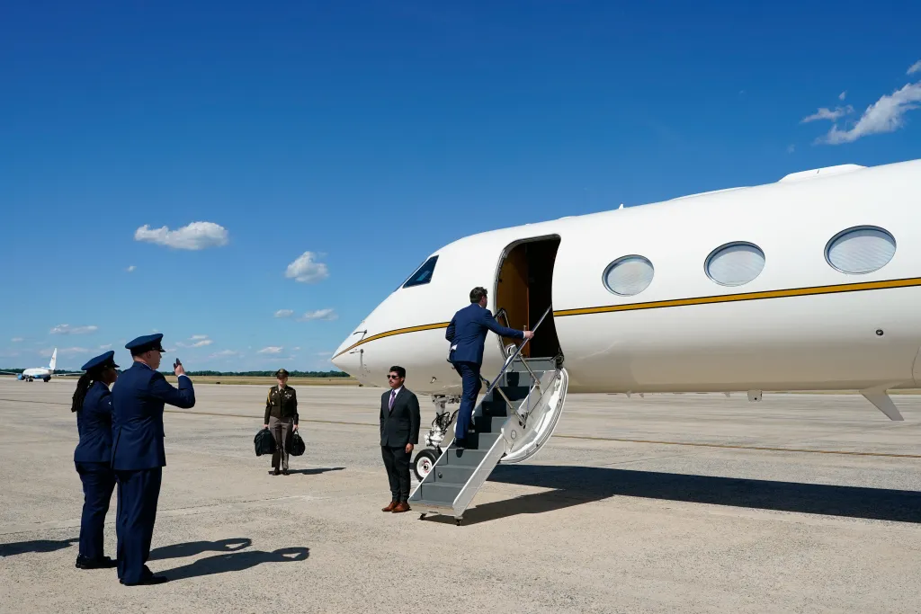 Man in a suit walking up the stairs to board a white private jet, with military personnel on the tarmac.