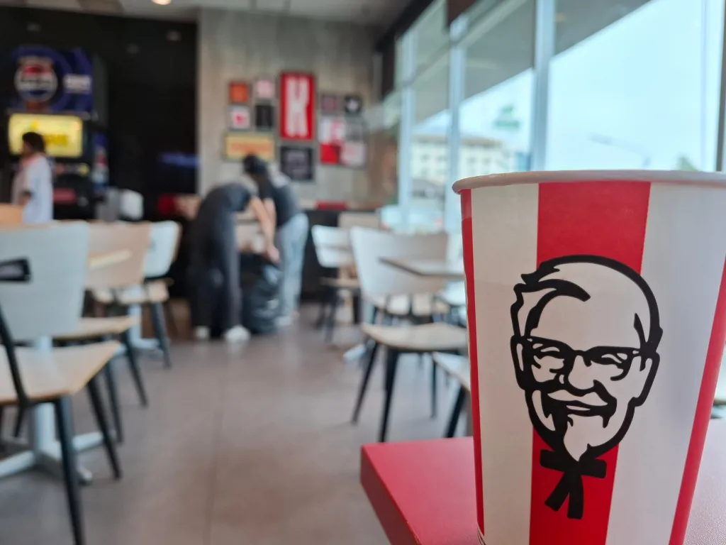 KFC cup with Colonel Sanders logo in the foreground of a blurred restaurant interior.