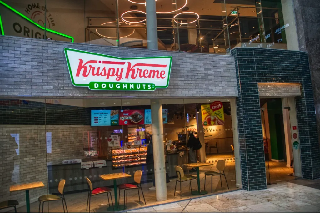 Krispy Kreme Doughnuts store with a large sign, display cases of doughnuts, and seating inside.