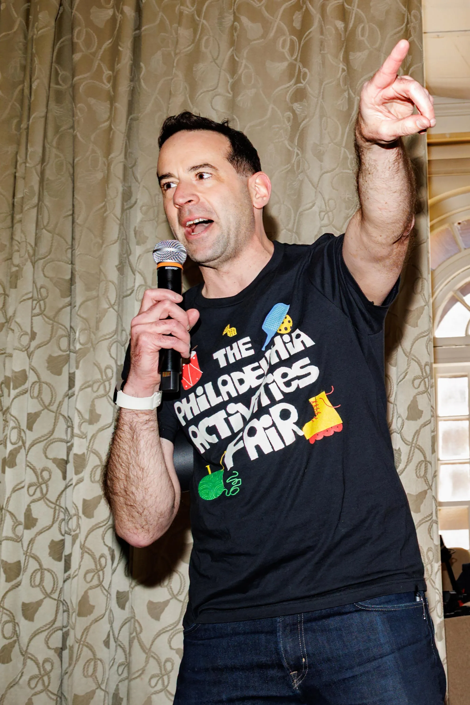 a man animatedly speaks into a microphone while pointing out of frame. His shirt reads “The Philadelphia Activities Fair”