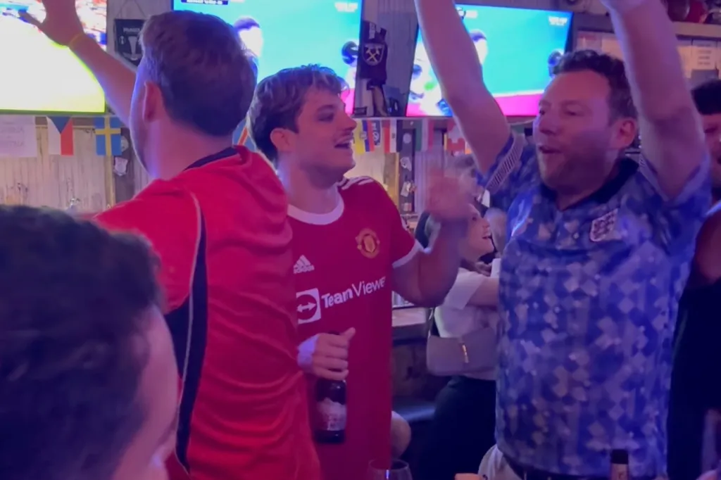 Spectators cheering with their arms in the air as they watch a soccer game on TV.