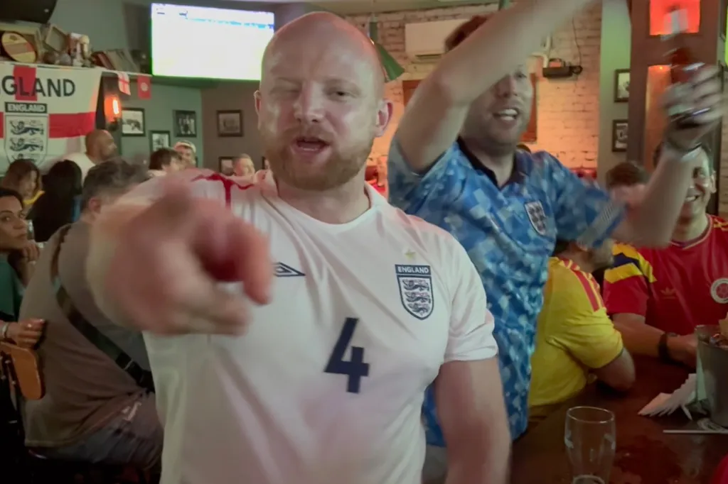 Man in an England soccer jersey pointing at the camera, celebrating in a crowded bar.
