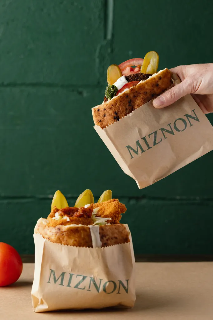 A hand holding a Miznon falafel pita sandwich over a Miznon fried chicken pita sandwich.