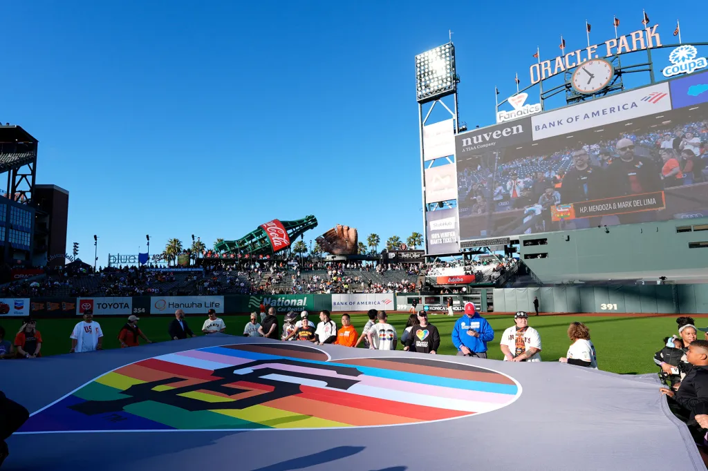 People on a baseball field holding a large San Francisco Giants logo banner, which is colored with the LGBTQ+ Progress Pride flag.