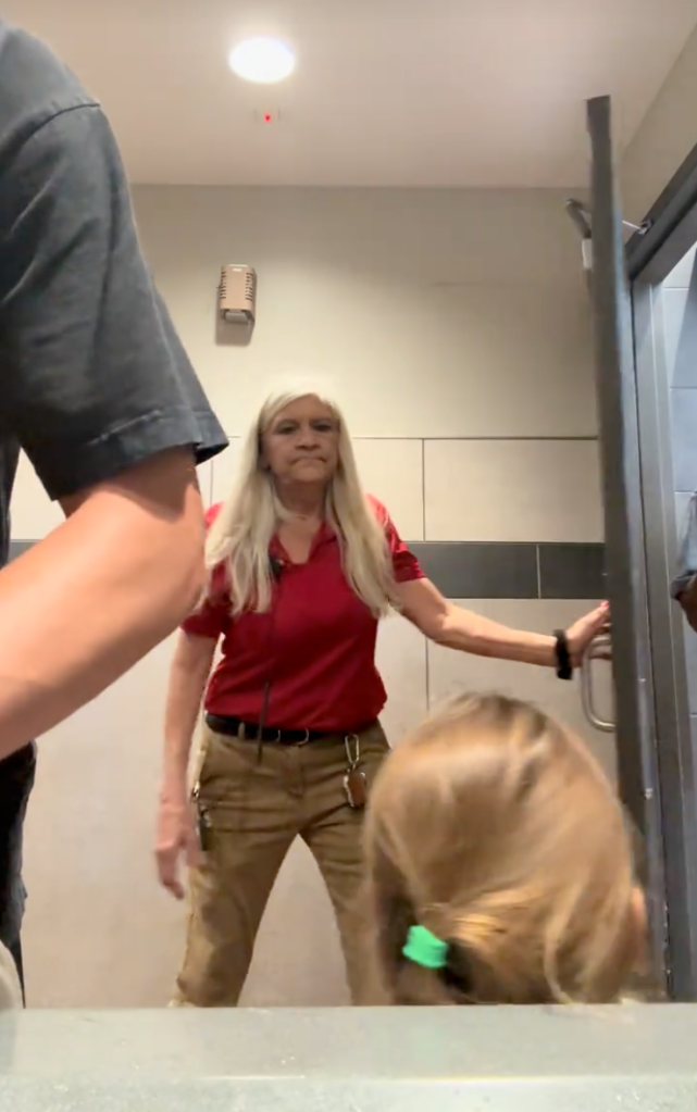 A gas station attendant in a red polo shirt, khaki pants, and long blonde hair looks at the camera while opening a door.