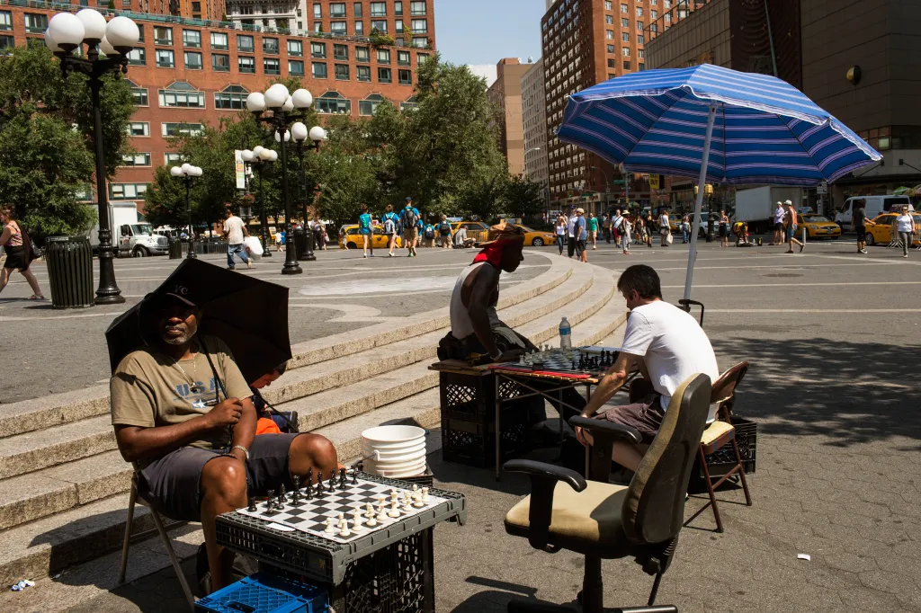 Two men play chess outdoors in a city square under umbrellas, while people walk by in the background.