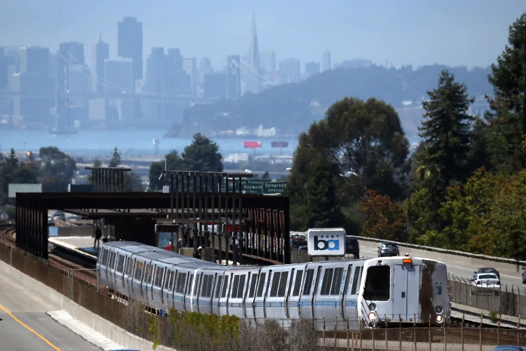 A BART train at a station with the San Francisco skyline and Bay Bridge in the hazy background.